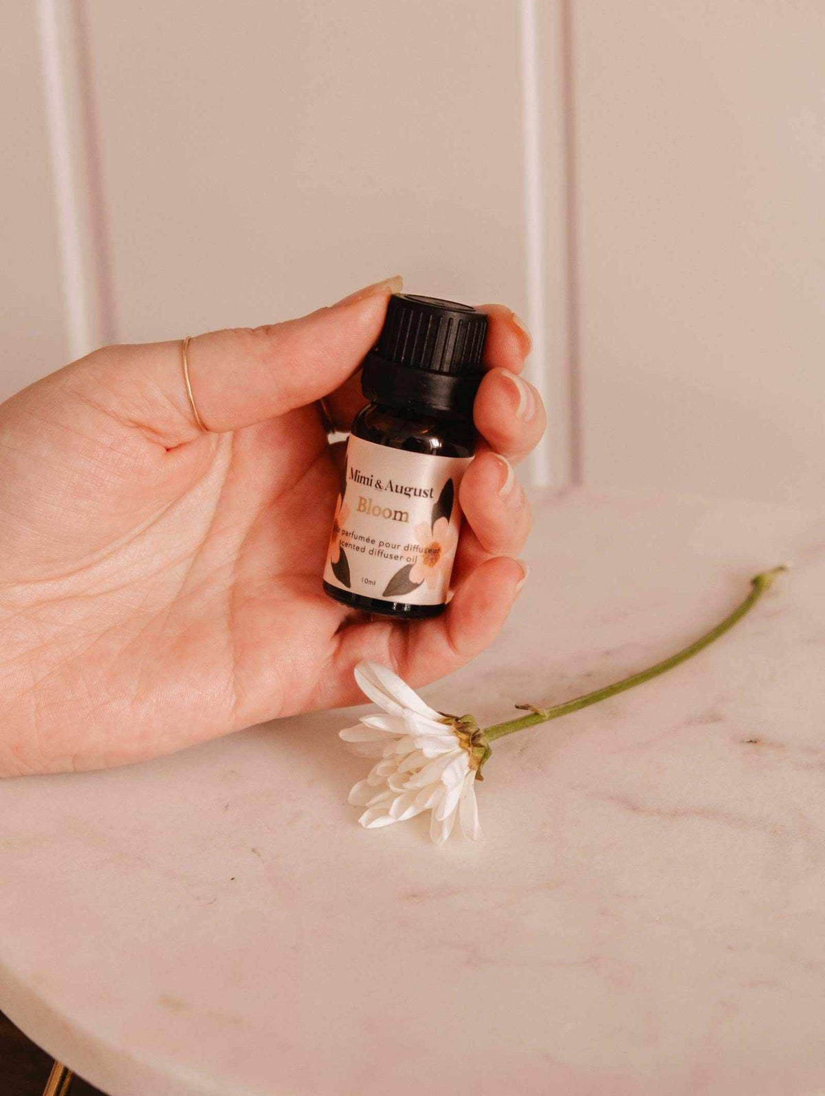A hand holds a small bottle labeled "Scented Oil - Bloom" from Mimi & August, releasing a refreshing fragrance beside a single white flower on a marble surface.