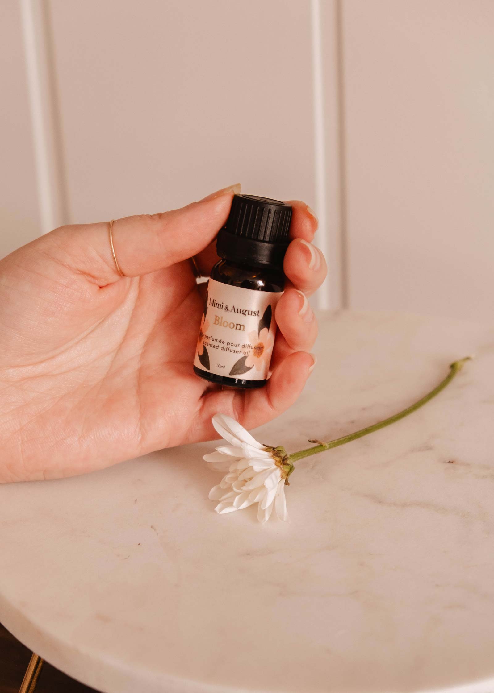 A hand holds a small bottle labeled "Scented Oil - Bloom" from Mimi & August, releasing a refreshing fragrance beside a single white flower on a marble surface.