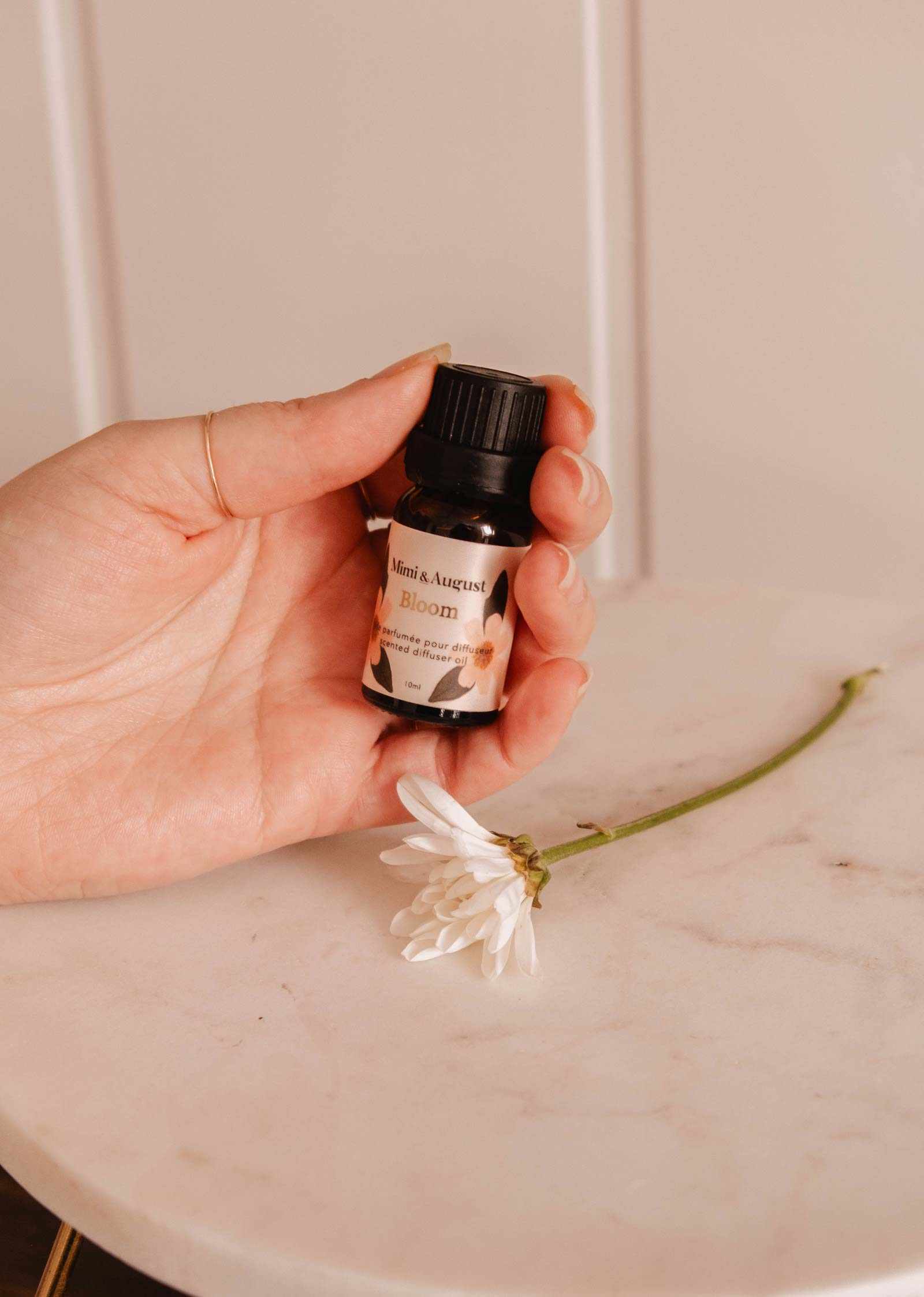Une main tient une petite bouteille étiquetée « Huile parfumée – Bloom » de Mimi & August, libérant une fragrance rafraîchissante à côté d'une fleur blanche sur une surface en marbre.