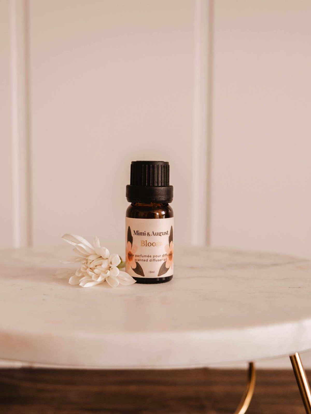 A small Scented Oil - Bloom by Mimi & August rests on a marble surface, releasing a refreshing fragrance with a small white flower beside it.