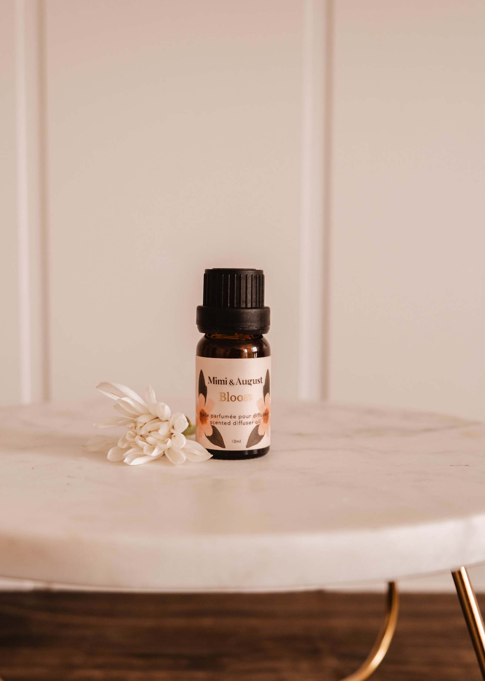 A small Scented Oil - Bloom by Mimi & August rests on a marble surface, releasing a refreshing fragrance with a small white flower beside it.