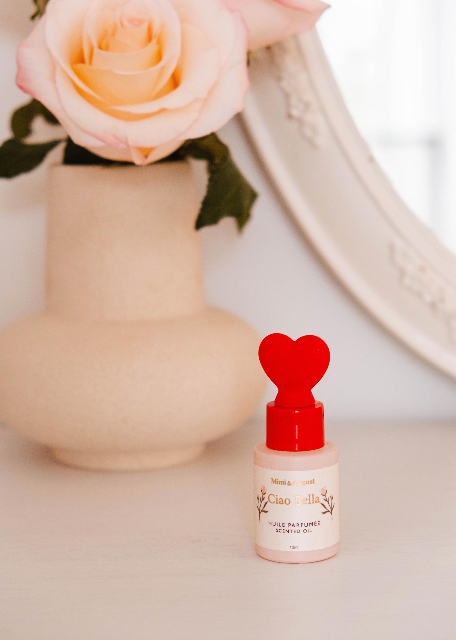 A bottle of Mimi & August's Scented Oil - Ciao Bella, with a red heart-shaped cap, sits before a beige vase holding a pink rose, releasing an elegant fragrance featuring jasmine and violet notes.