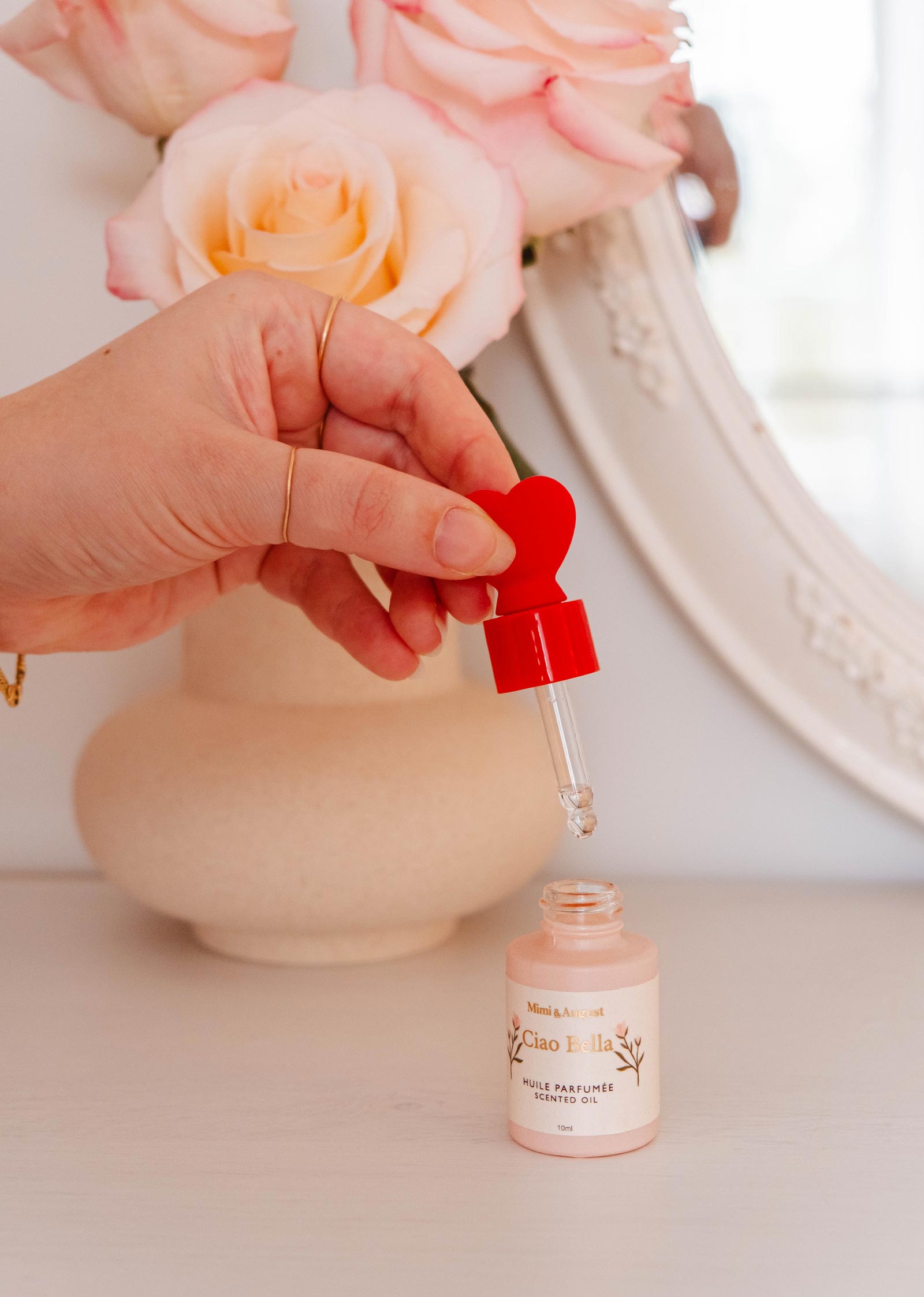 A hand delicately holds a red dropper above a small bottle labeled "Ciao Bella" by Mimi & August, releasing an elegant fragrance of jasmine, violet, cedar, and citrus. A vase of pink roses and a mirrored backdrop complete this enchanting scene.