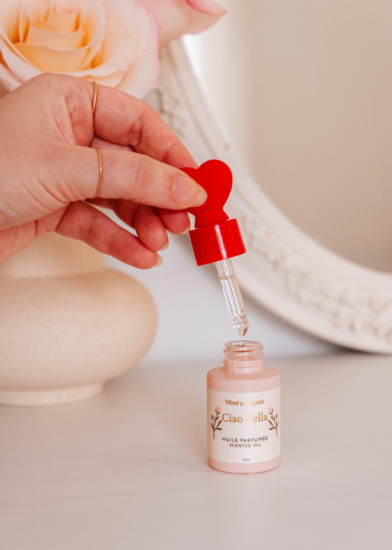 A hand holds a dropper above a small bottle labeled "Scented Oil - Ciao Bella" by Mimi & August, with a red heart-shaped top. A rose and decorative mirror in the background create an elegant ambiance with jasmine, violet, cedar, and citrus notes.