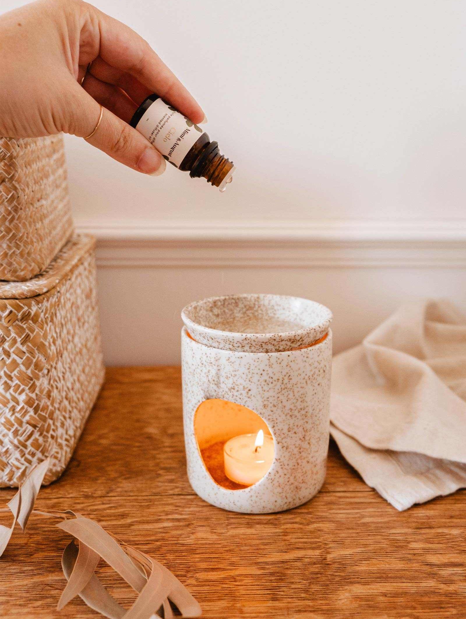 A hand is pouring Mimi & August's Scented Oil - Cielo from a bottle into a ceramic diffuser with a lit tea light candle inside, releasing a calming scent of warm amber. Woven baskets and a beige cloth rest on the wooden surface next to the diffuser.