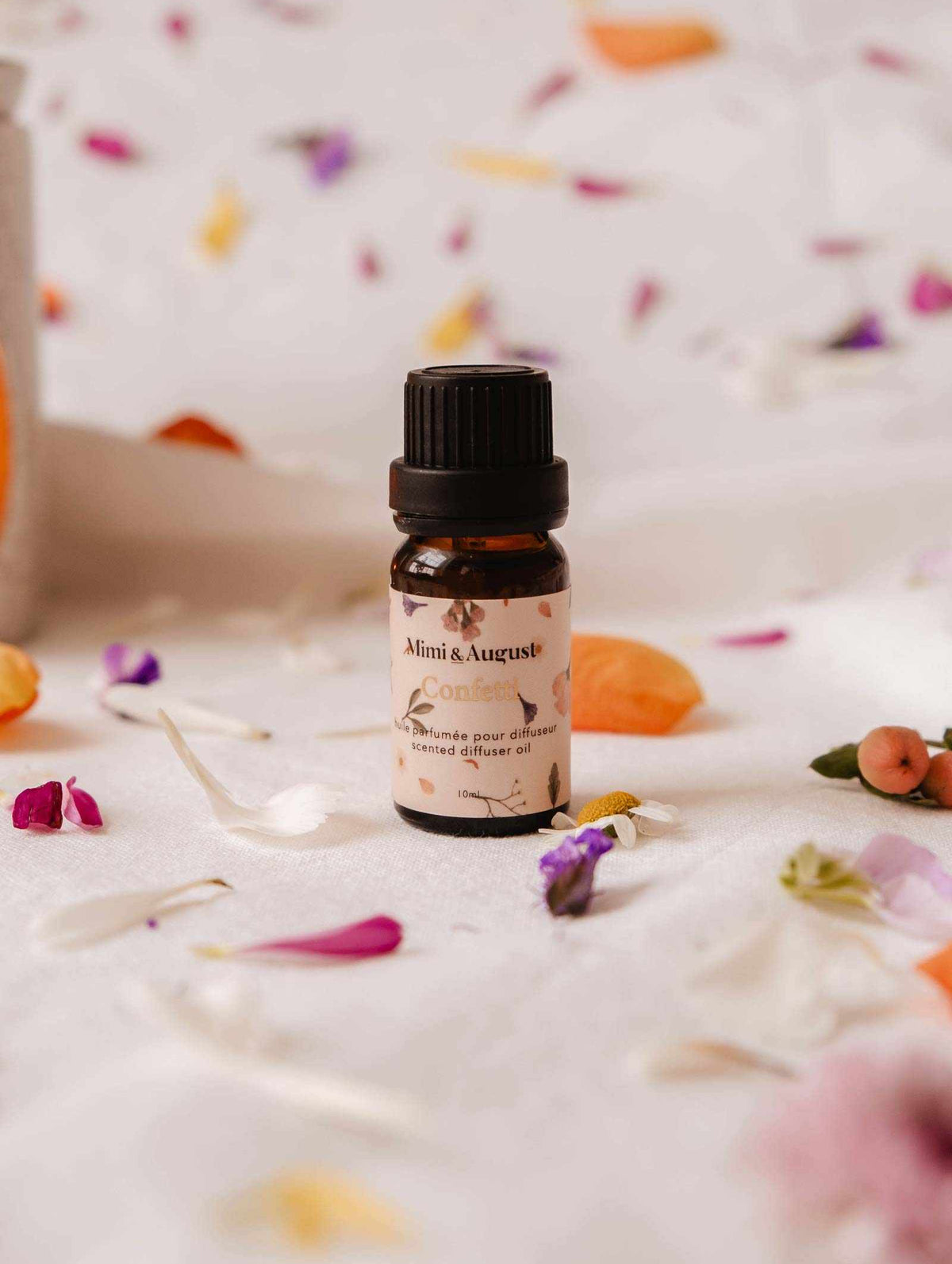 A small amber bottle labeled "Mimi & August Confetti" rests on a white fabric surface, surrounded by scattered colorful flower petals. The Scented Oil - Confetti fills the air with subtle mandarin notes.
