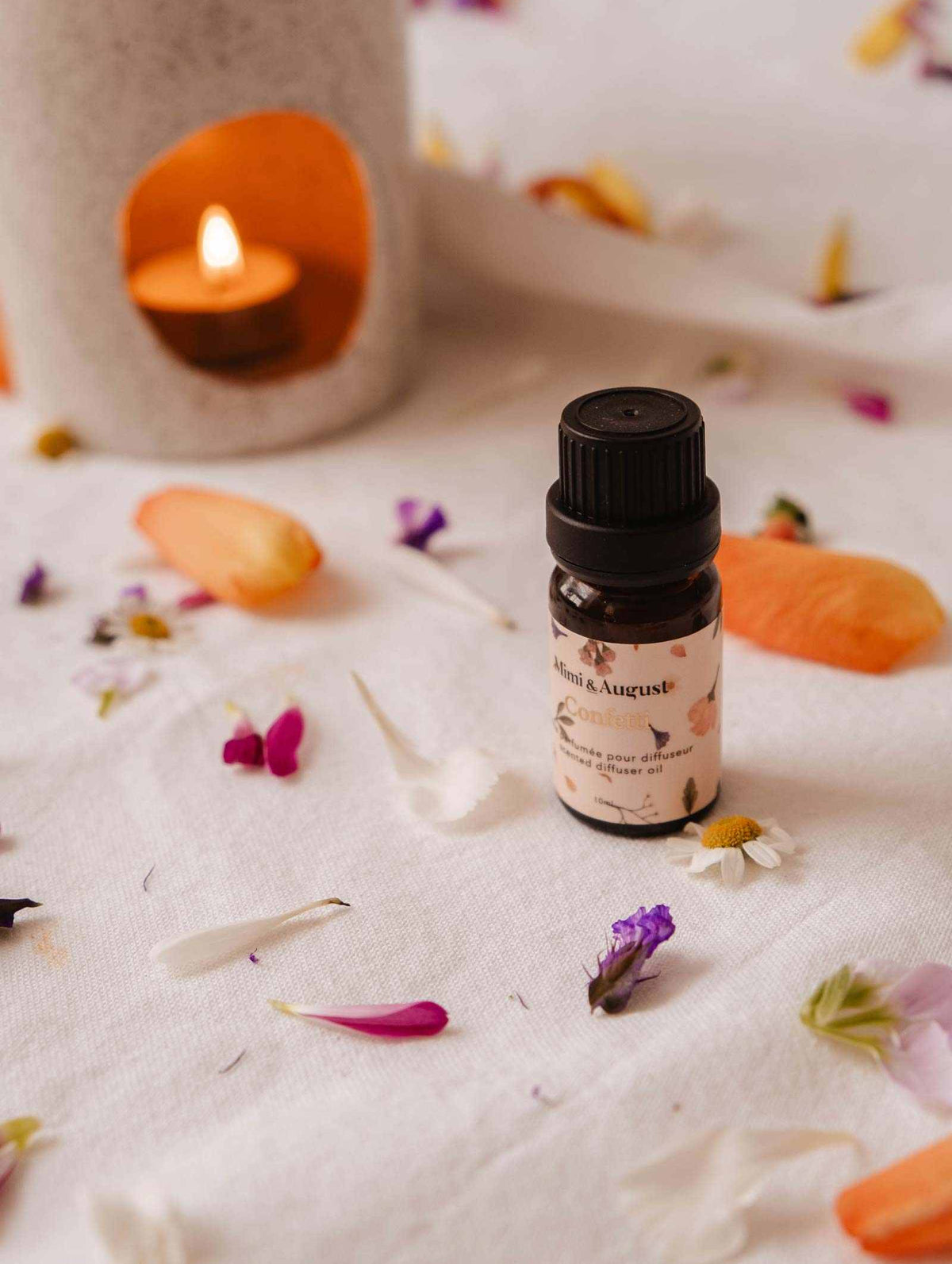A small bottle of Mimi & August's Scented Oil - Confetti sits on a white cloth with scattered flower petals, releasing its fragrance. A lit candle flickers softly in the background, adding warmth and tranquility to the scene.