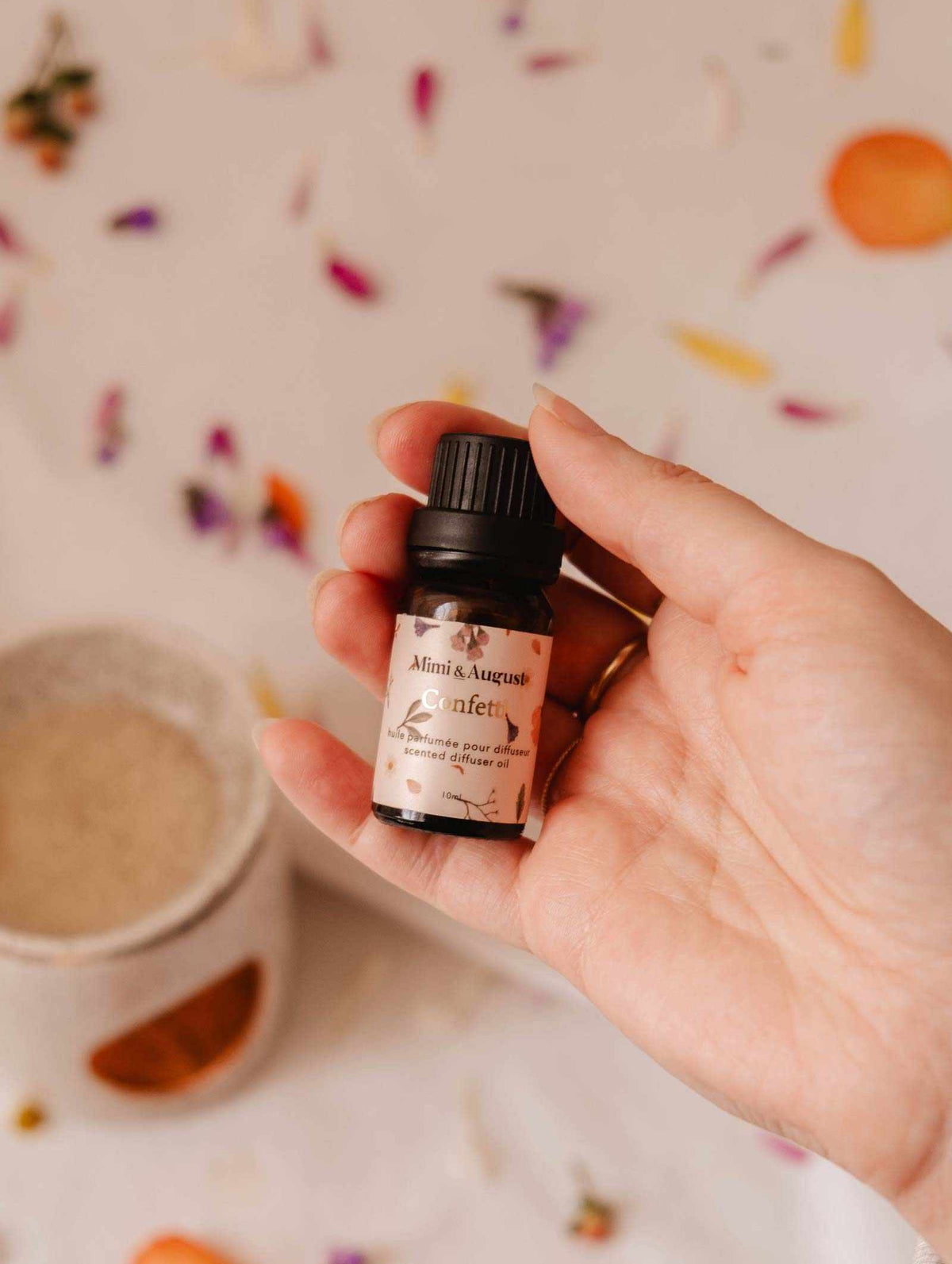 A hand holds a small bottle of Mimi & August's "Scented Oil - Confetti" amidst scattered flower petals and a ceramic oil burner, suggesting a fragrant aroma with hints of mandarin.