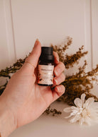 A hand holds a small bottle of "Fleurette" scented oil by Mimi & August, evoking wildflower fields, with dried flowers in the background.