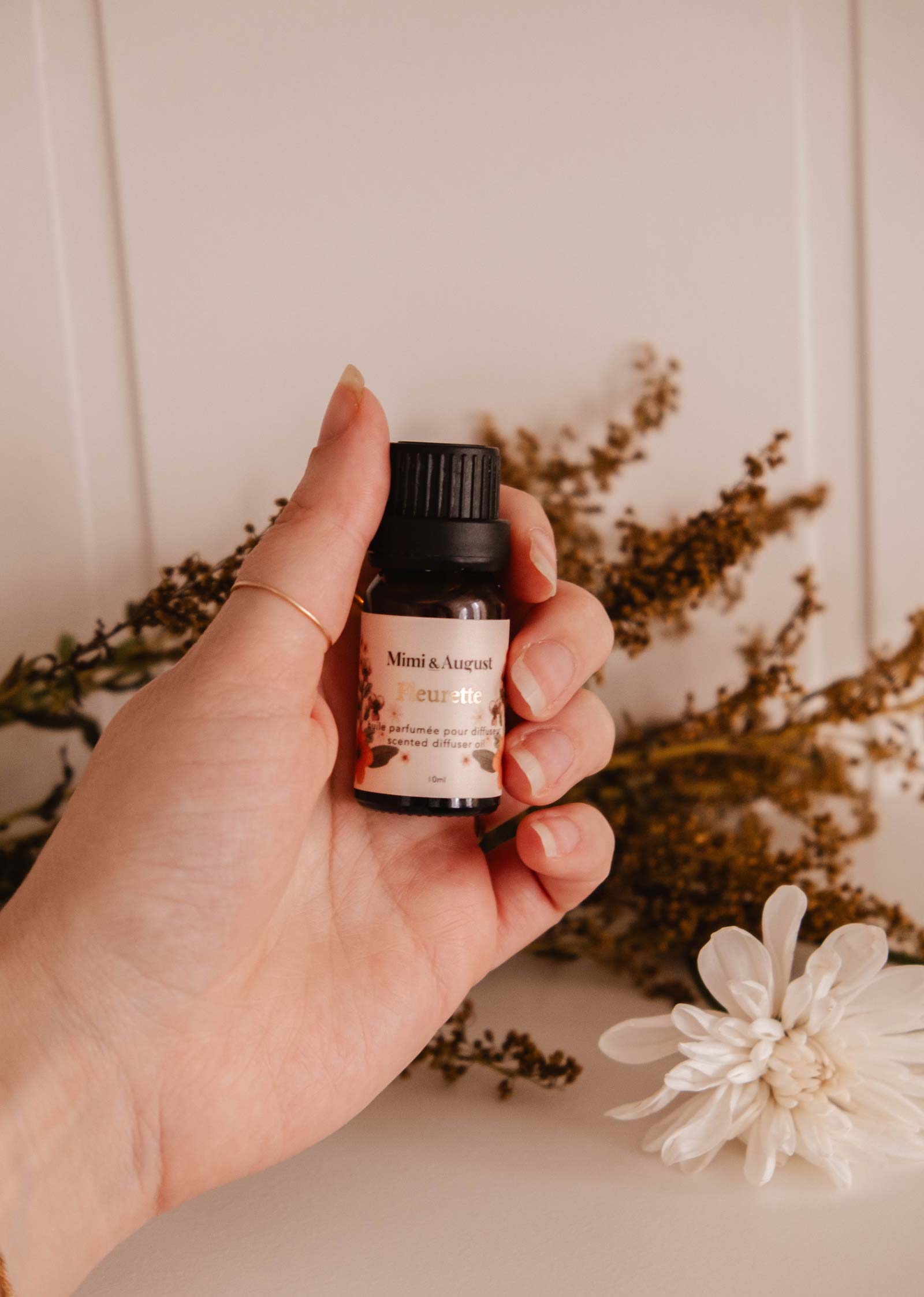 A hand holds a small bottle of "Fleurette" scented oil by Mimi & August, evoking wildflower fields, with dried flowers in the background.