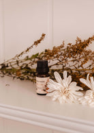 A small bottle of Mimi & August's Scented Oil - Fleurette with a label is placed on a white surface, surrounded by dried herbs and two white flowers, exuding delicate floral scents.