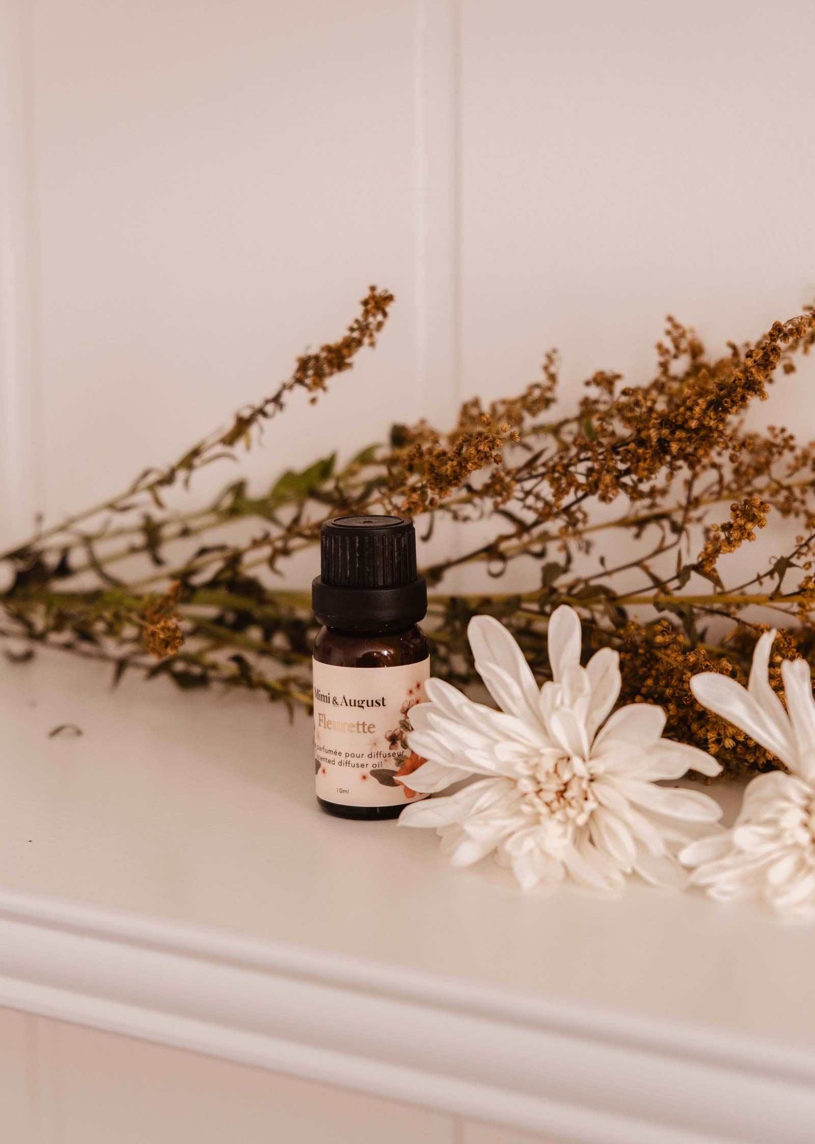 A small bottle of Mimi & August's Scented Oil - Fleurette with a label is placed on a white surface, surrounded by dried herbs and two white flowers, exuding delicate floral scents.