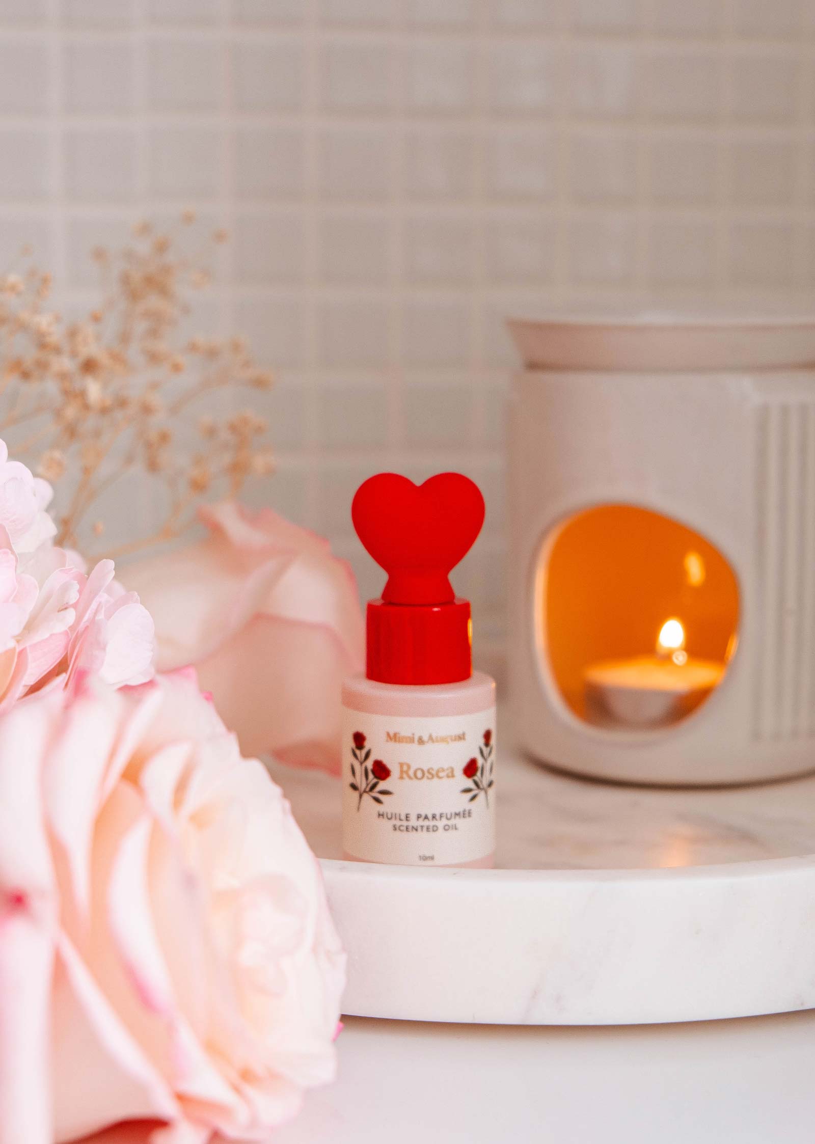A Mimi & August Scented Oil - Rosea bottle with a heart-shaped cap sits on a marble tray next to pink flowers, releasing a citrus and rose aroma. A lit candle flickers warmly in the background.