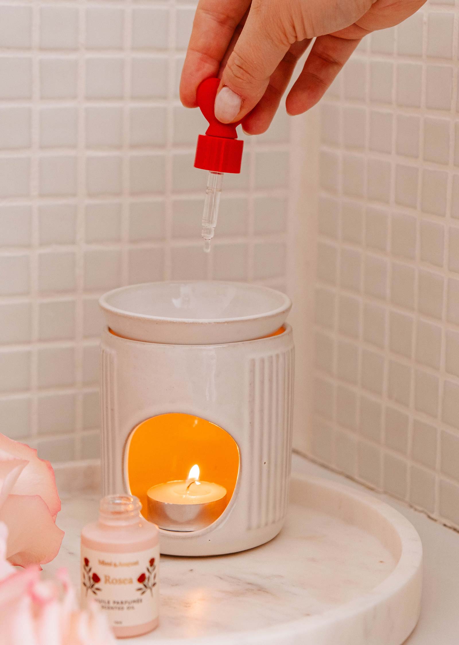 A hand adds drops of Mimi & August's Scented Oil - Rosea from a red dropper into a white diffuser, releasing a citrus and rose aroma. A lit tealight candle inside casts a warm glow on the tiles.