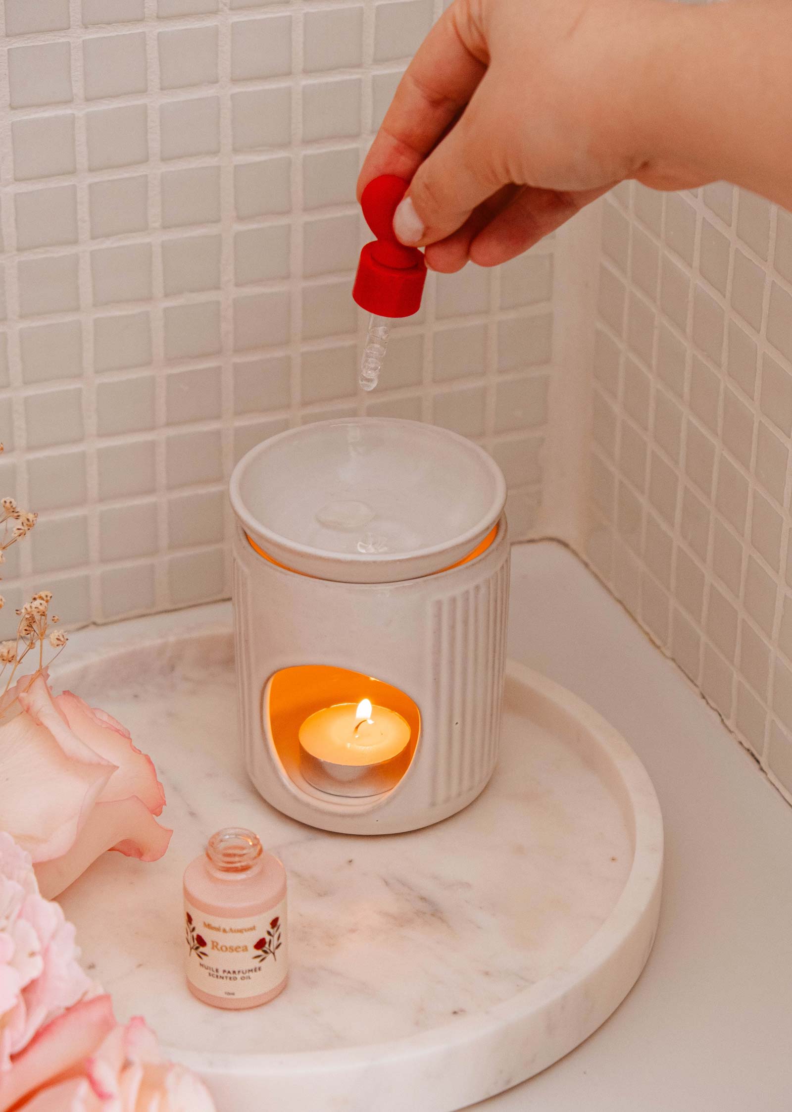 A hand uses a dropper to add Mimi & August's Scented Oil - Rosea to a ceramic wax warmer with a lit tealight. The bottle sits on a marble tray beside pink roses, filling the air with its delicate aroma.