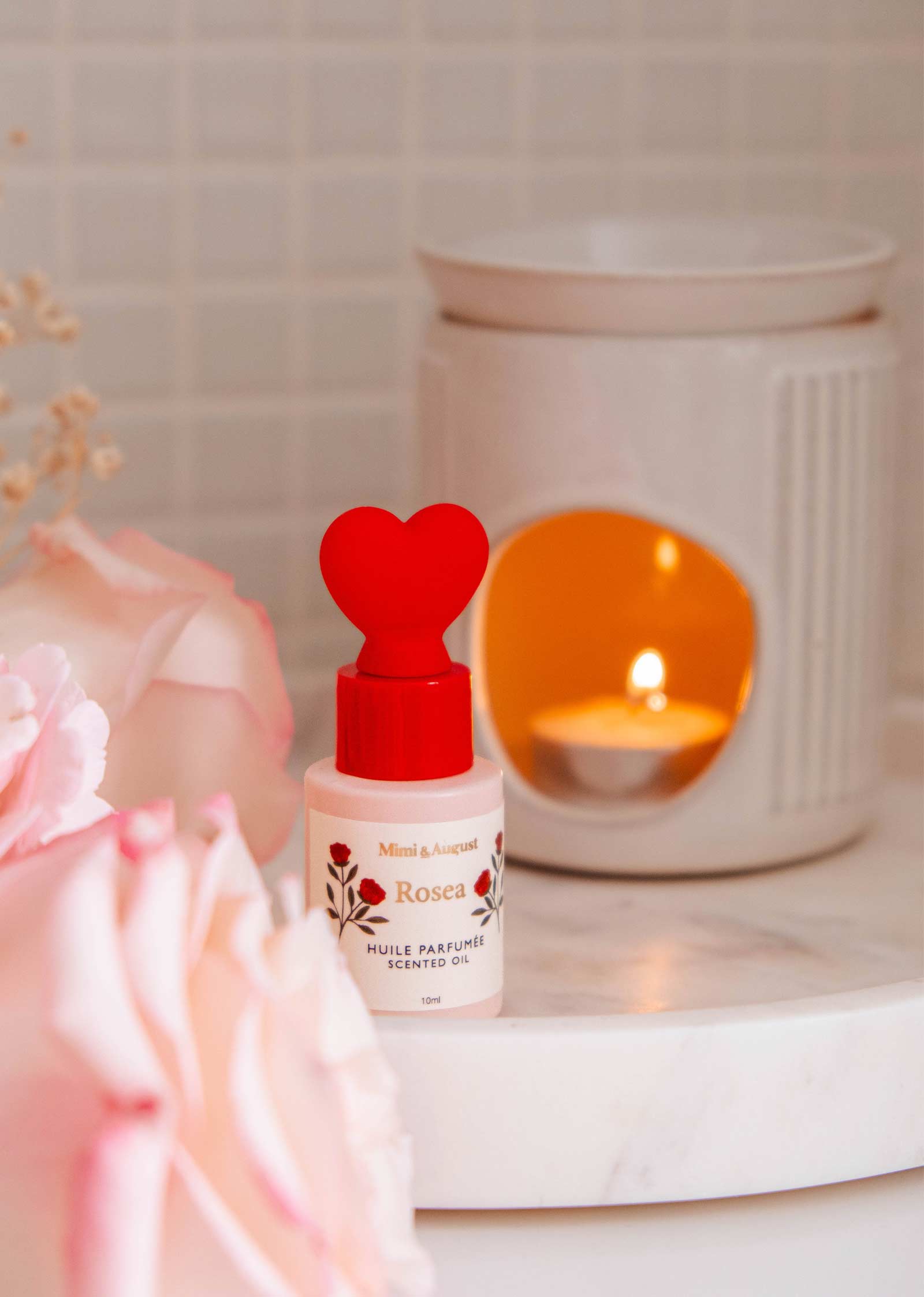 A Mimi & August Rosea scented oil bottle with a red heart cap sits next to pink roses and a lit candle on white marble, filling the air with citrus and rose scents.