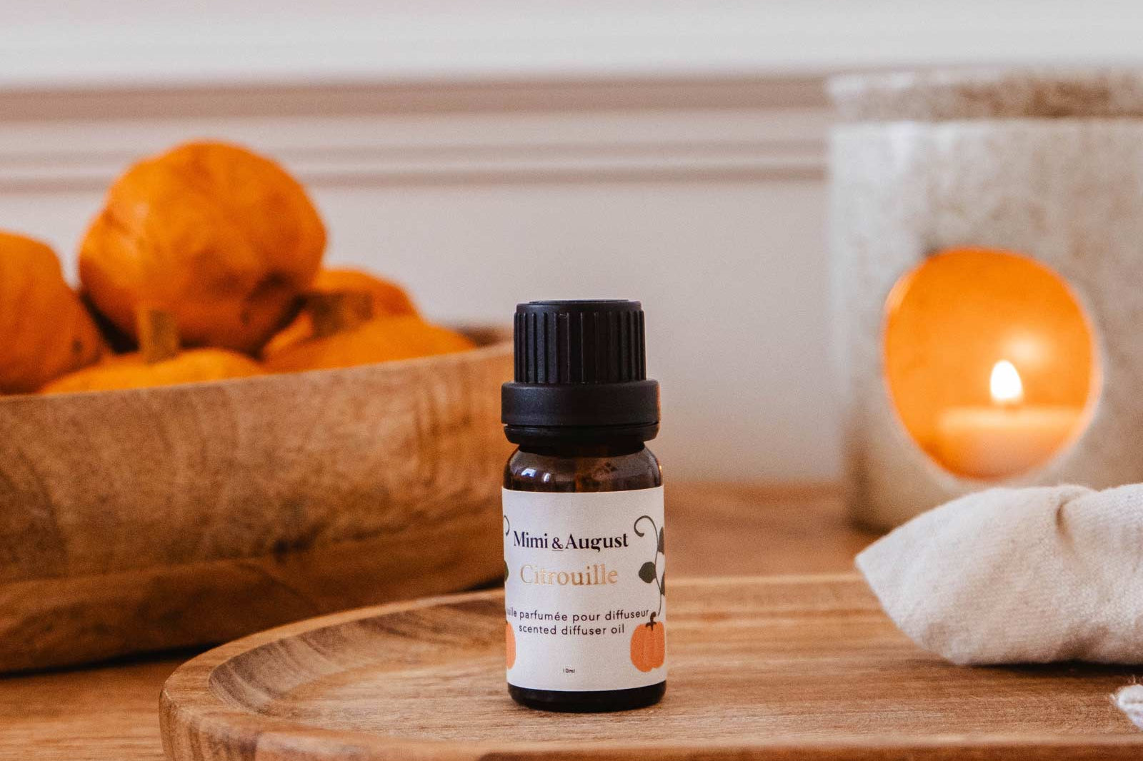 A small bottle of Mimi & August Scented Oil - Citrouille is placed on a wooden tray, with a candle and bowl of orange items in the background, perfect for relaxation when used in your diffuser.