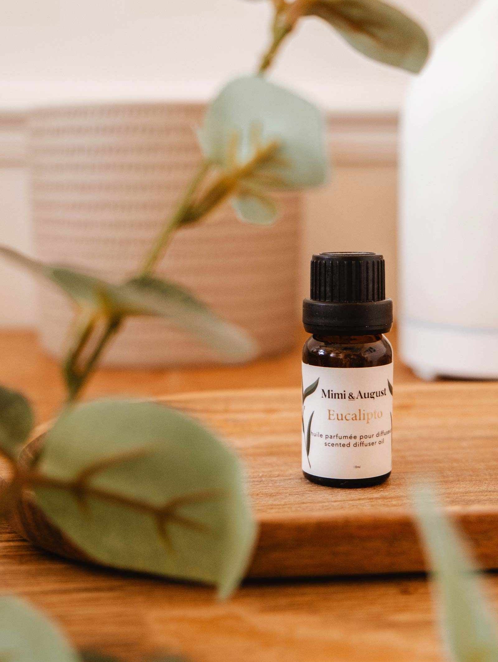 A small bottle labeled "Scented Oil - Eucalipto" from Mimi & August is placed on a wooden surface, surrounded by leaves. The eucalyptus scent offers a relaxing and refreshing atmosphere.