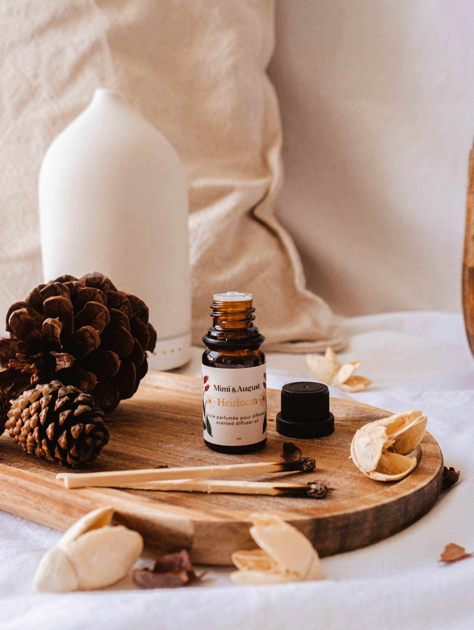 A small bottle of Heirloom Scented Oil by Mimi & August is displayed on a wooden tray adorned with pine cones, dried leaves, and some wood sticks. An aromatherapy diffuser in the background completes the perfect relaxation escape.