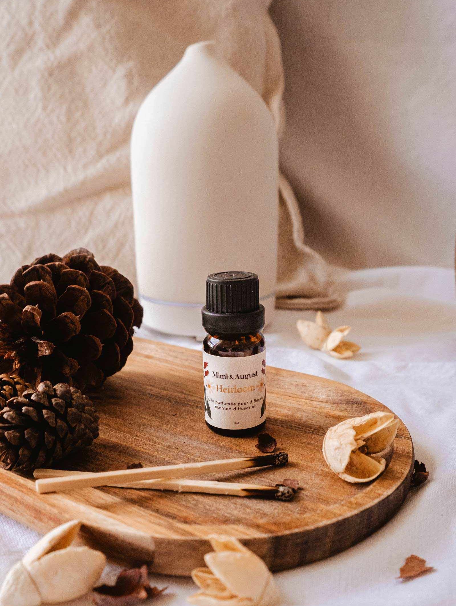 A small bottle of Mimi & August's Scented Oil - Heirloom is placed on a wooden tray next to pinecones, wooden sticks, and a white vase, offering a touch of relaxation and escape.
