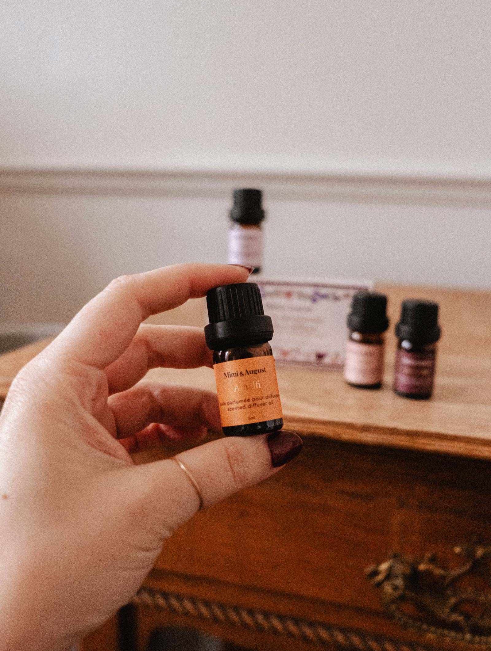 A hand holds a small amber bottle labeled “Scented Oils Discovery Kit” by Mimi & August, with more small bottles and boxes from the collection arranged on a wooden table in the background.