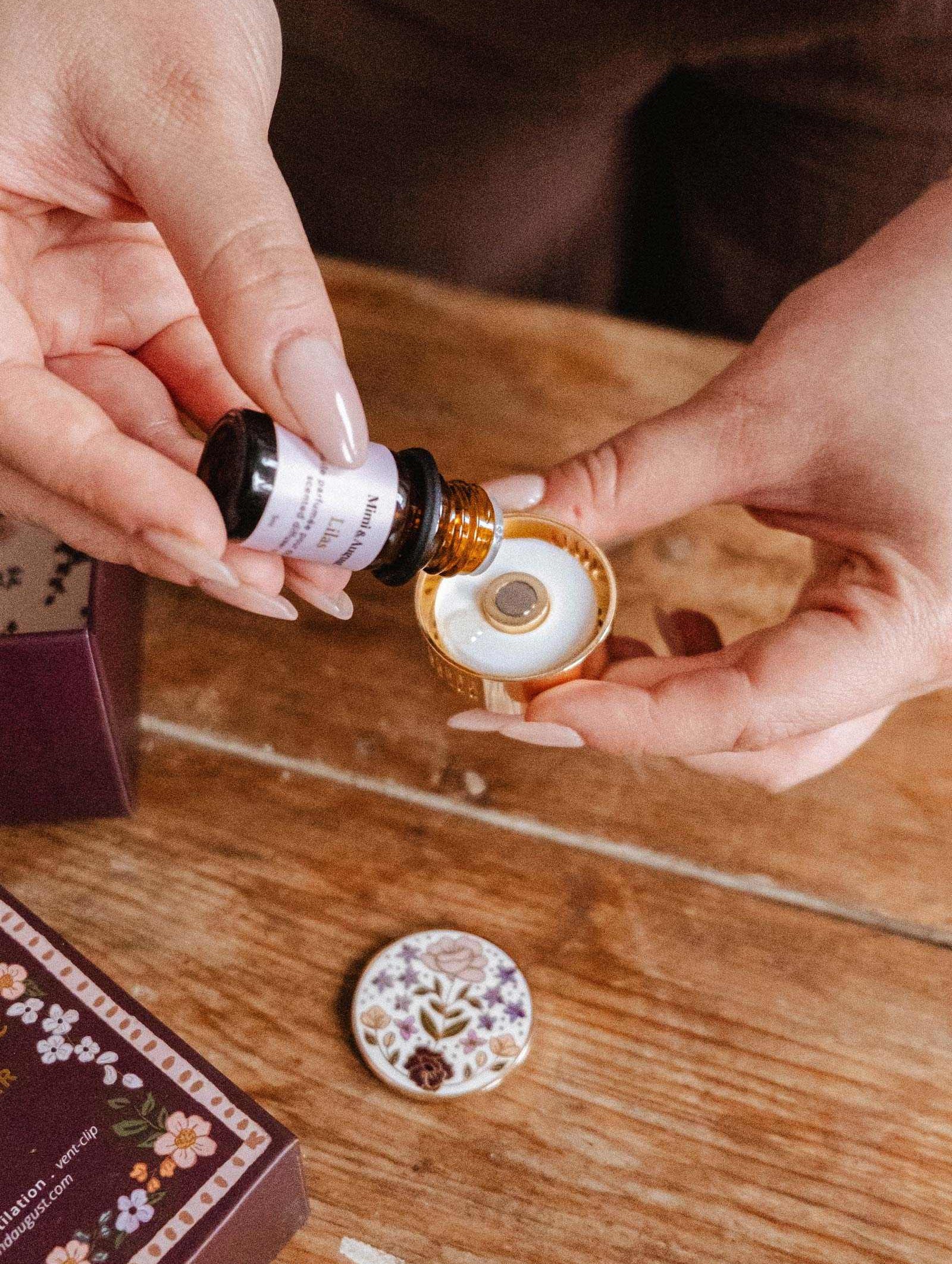 Someone explores their fragrance collection using the Mimi & August Scented Oils Discovery Kit, pouring oils from a small bottle into a round ceramic diffuser with a floral-patterned lid on a wooden table.
