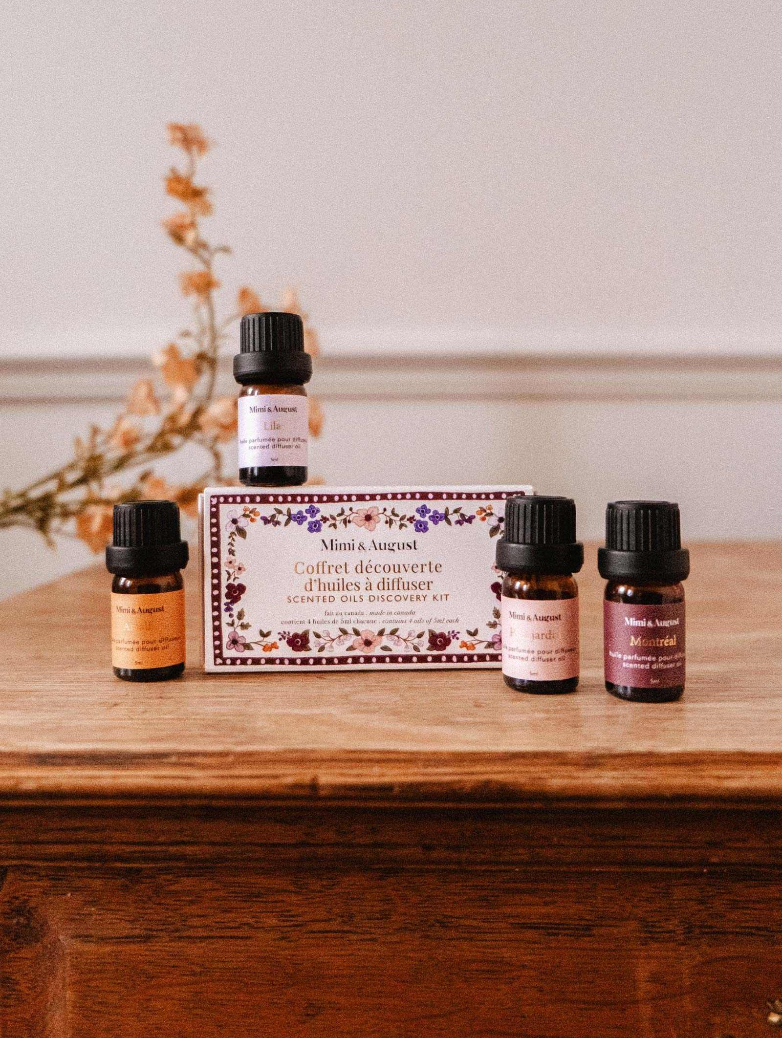 The Scented Oils Discovery Kit by Mimi & August, featuring four small bottles in a labeled box, is displayed on a wooden surface with dried flowers in the background.