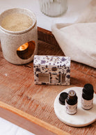 A ceramic holder with a lit candle, a floral design box, and Mimi & August's Secret Garden Scented Oil Trio adorn the wooden surface, crafting an inviting ambiance.