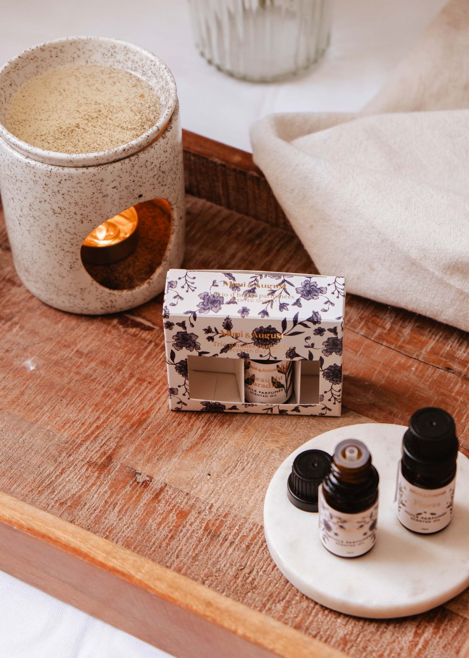 A ceramic holder with a lit candle, a floral design box, and Mimi & August's Secret Garden Scented Oil Trio adorn the wooden surface, crafting an inviting ambiance.