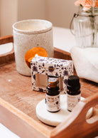 A wooden tray contains a packaged Secret Garden Scented Oil Trio by Mimi & August, two small bottles, a ceramic candle holder, and a glass jar with flowers.