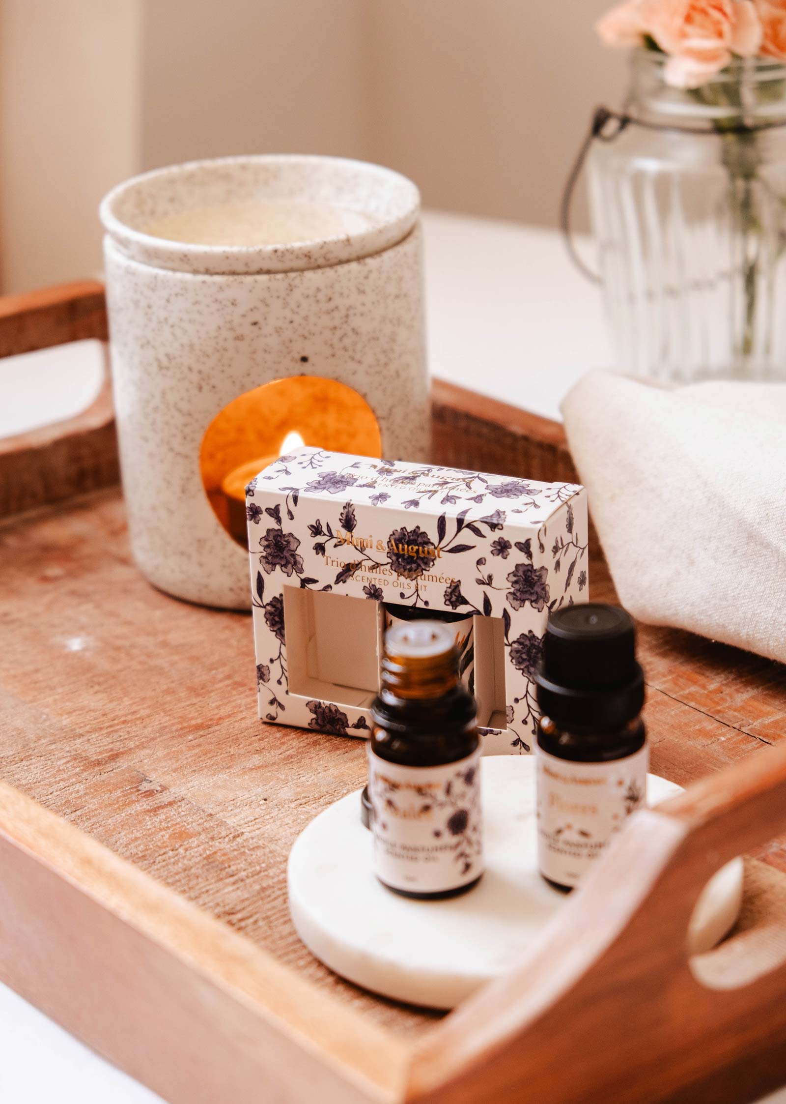 A wooden tray contains a packaged Secret Garden Scented Oil Trio by Mimi & August, two small bottles, a ceramic candle holder, and a glass jar with flowers.