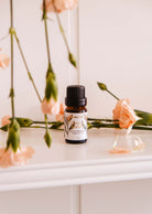 A bottle from the Mimi & August "Secret Garden Scented Oil Trio" set exudes a tropical fragrance as it rests on a white surface, surrounded by pink flowers, evoking floral charm.