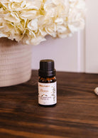 A small bottle from the Mimi & August "Secret Garden Scented Oil Trio" sits on a wooden surface, emanating a tropical aroma. Nearby, a wicker basket overflowing with white hydrangeas adds to the serene floral ambiance.