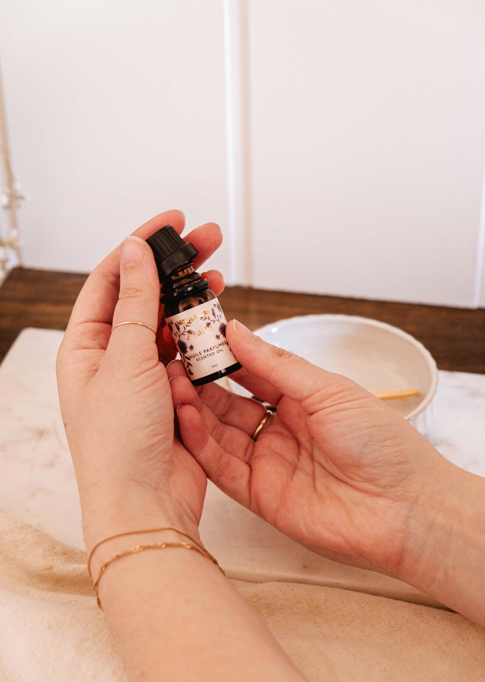 Hands holding a small dropper bottle with a floral charm label, above a table next to a white bowl with a wooden stick. Mimi & August's Set | Secret Garden Scented Oil Trio enchants with tropical fragrances that transport you to lush, exotic landscapes.