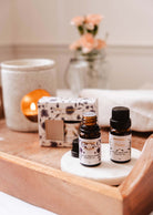 The Secret Garden Scented Oil Trio by Mimi & August includes two essential oil bottles on a wooden tray with a decorative box, candle, and towel. A floral vase enhances the fragrance, creating an inviting and serene atmosphere.