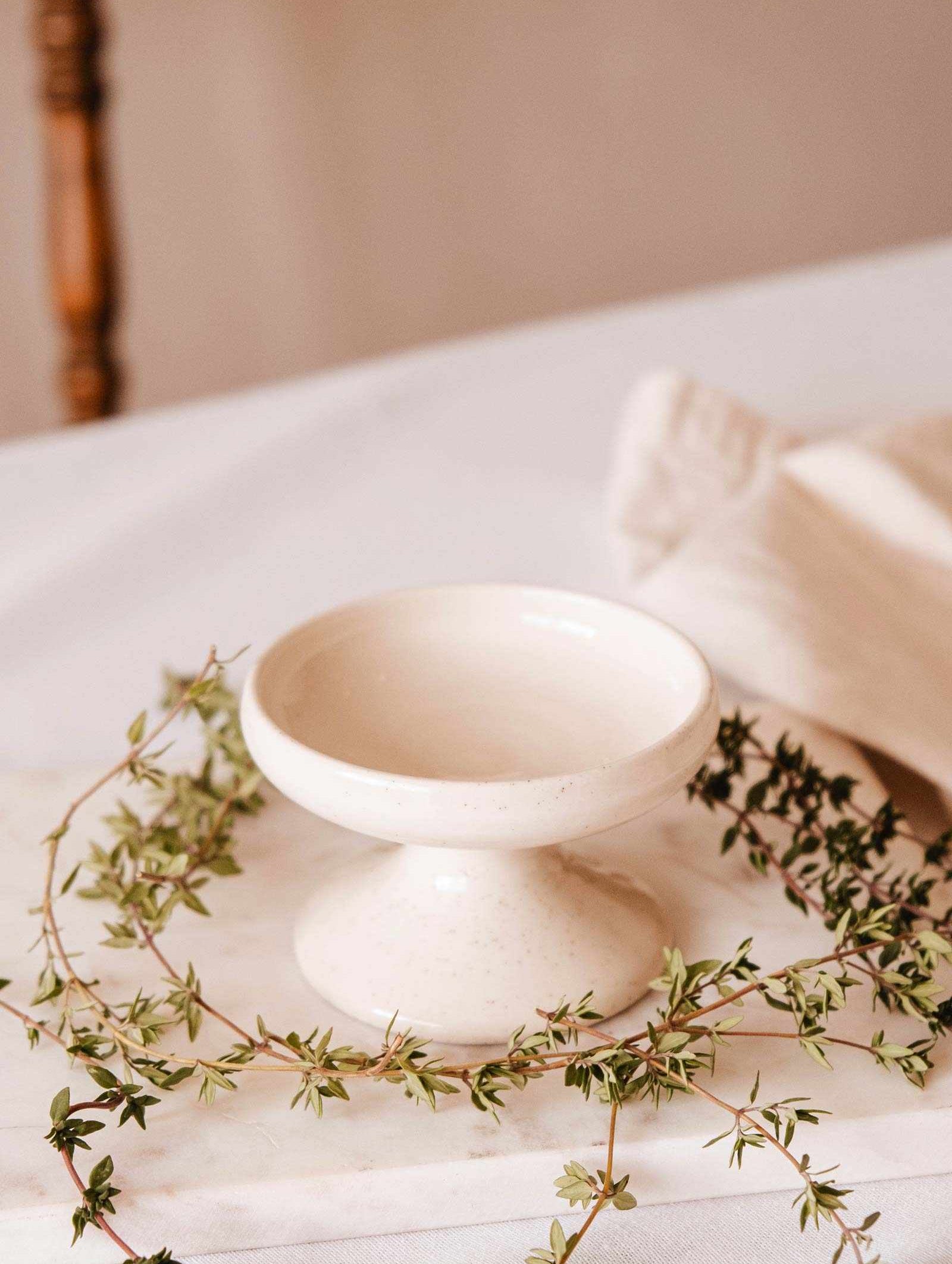 A handcrafted ceramic pedestal bowl rests on a marble surface, surrounded by greenery and a folded beige cloth, exuding the tranquil charm of the Serenity Pillar Candle Holder by Mimi & August.