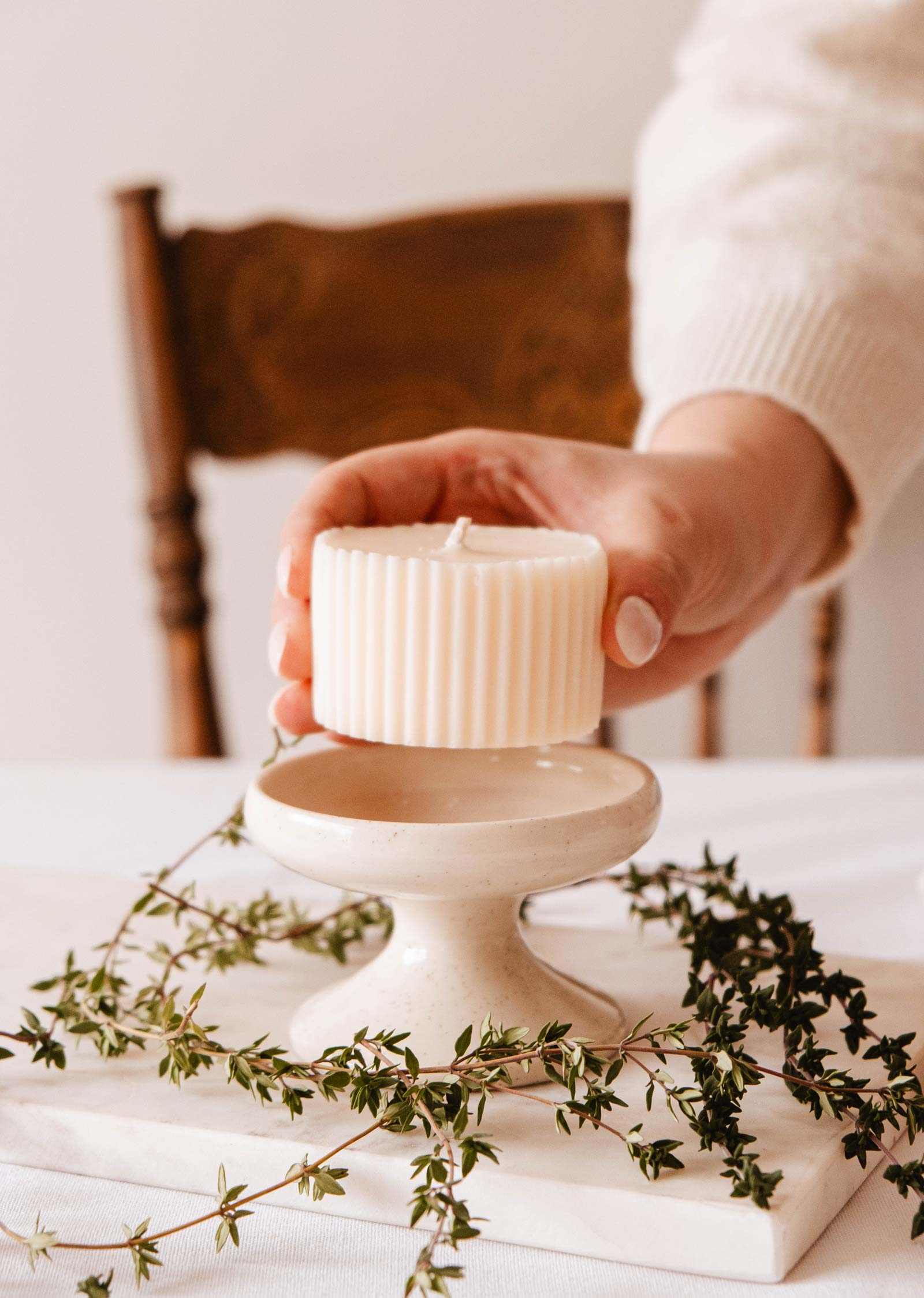 A hand places a ribbed, cream-colored Mimi & August fig candle refill on a ceramic stand, surrounded by green leaves and a wooden chair, filling the air with its fig cassis amber scent.