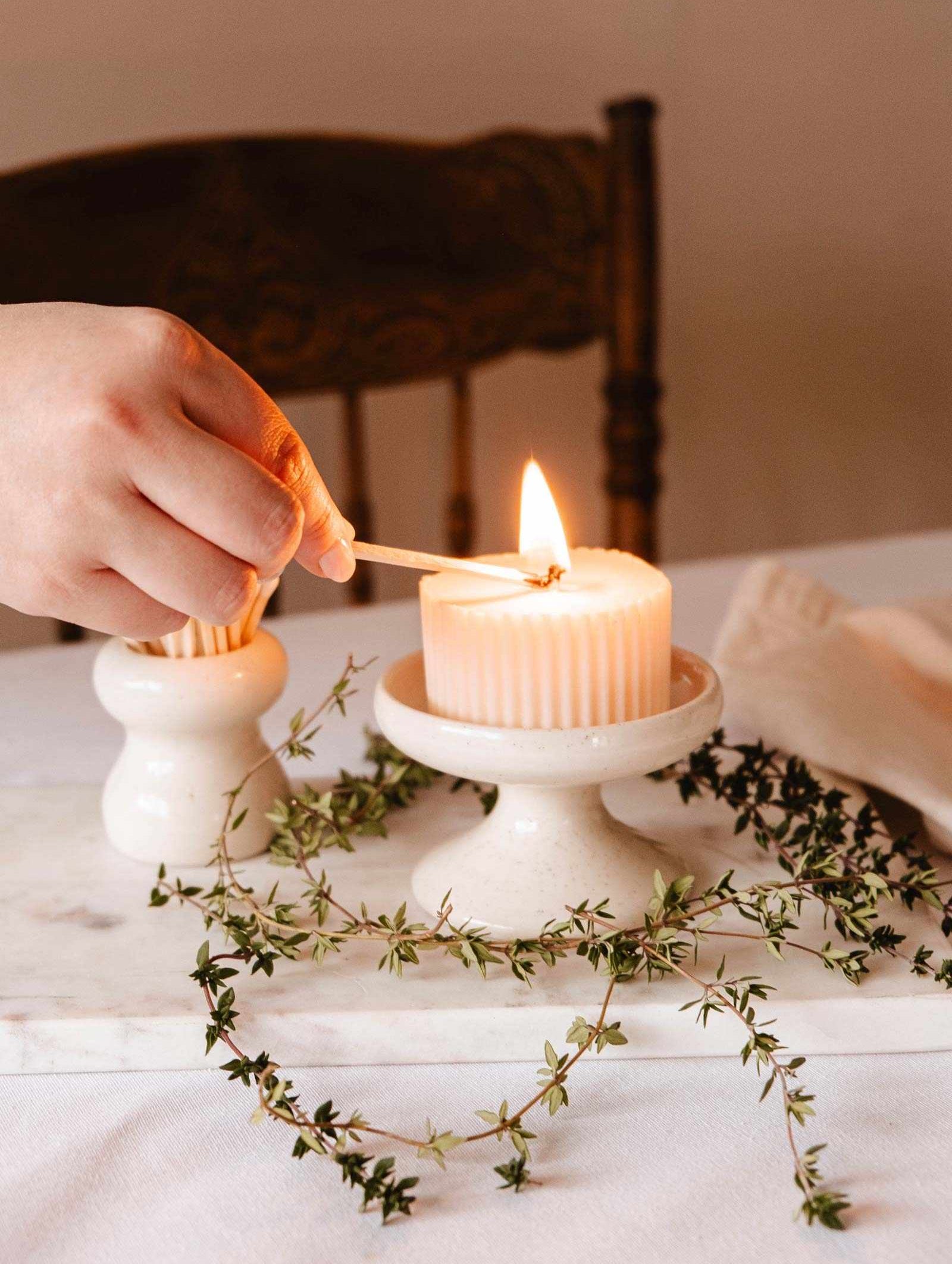 A hand lights a ribbed candle on the Mimi & August Serenity Pillar Candle Holder, which is on a marble stand with green twigs.