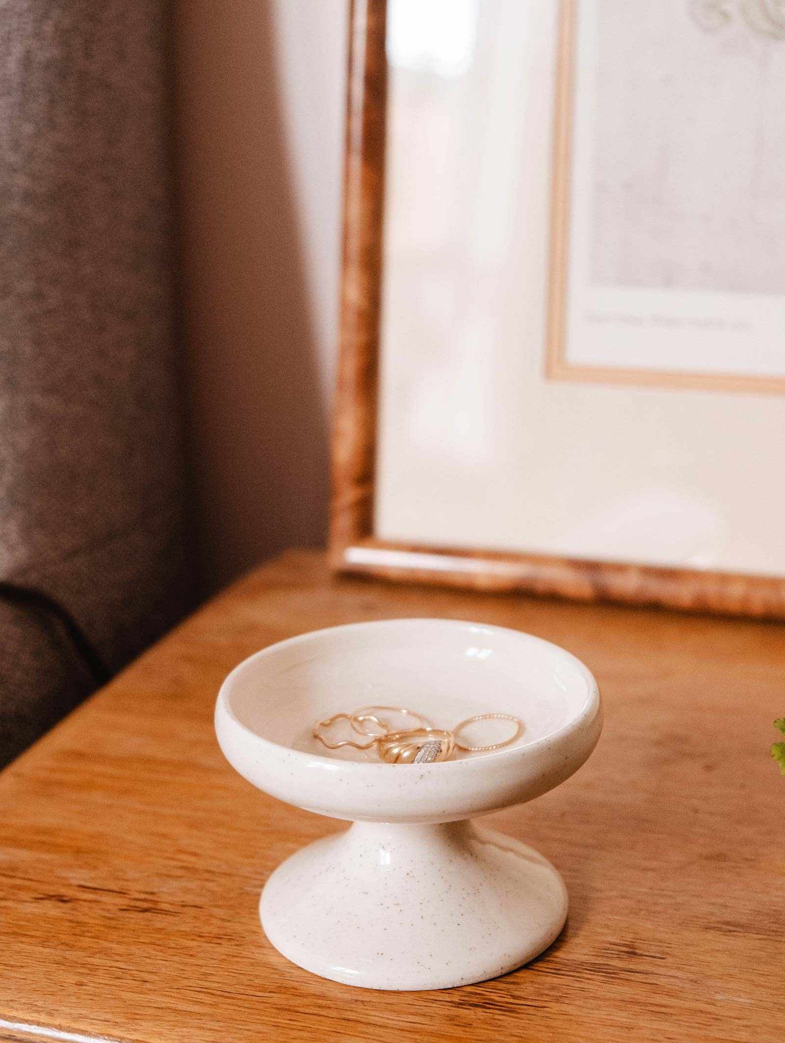 A Serenity Pillar Candle Holder by Mimi & August sits on a wooden surface holding several pieces of jewelry, while a partially visible framed picture enhances the versatile home decor.