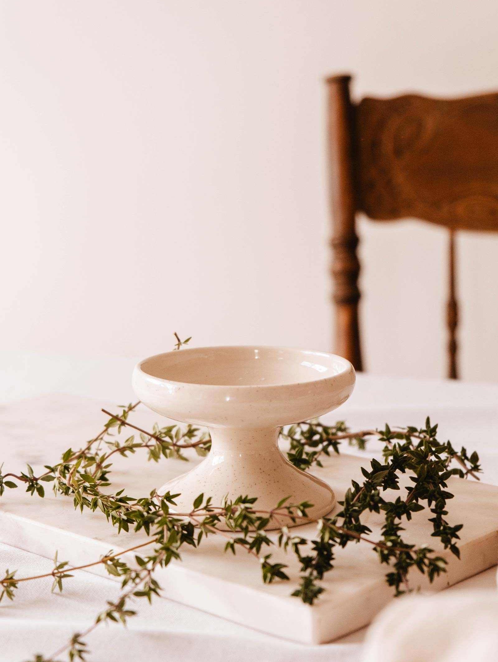 The Serenity Pillar Candle Holder by Mimi & August rests on a marble slab adorned with green vines, next to a wooden chair.