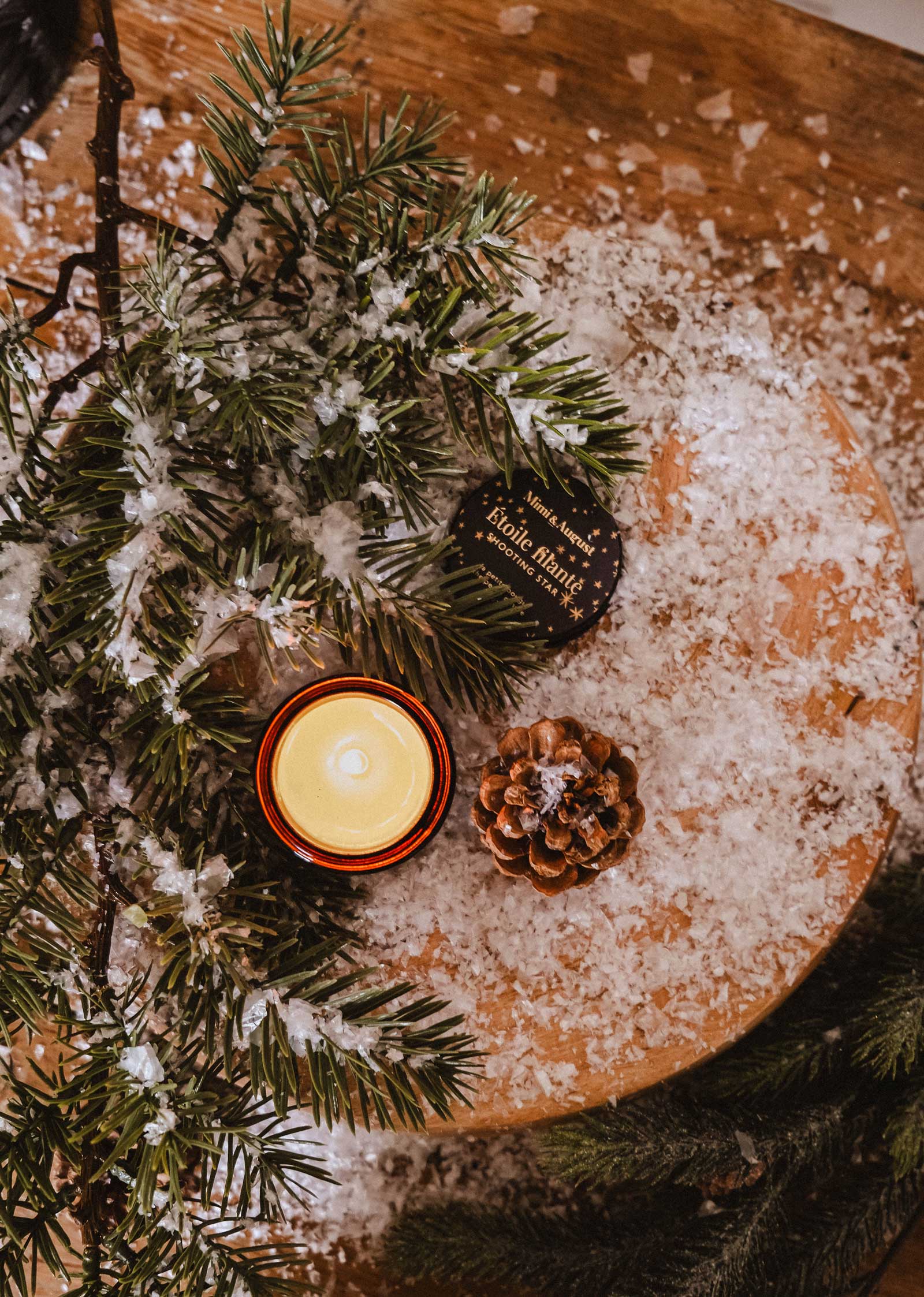 A festive Mimi & August Shooting Star 2 oz candle, pinecone, and fir branches on a wooden snowy surface beside a small “Exotic Forest” label.
