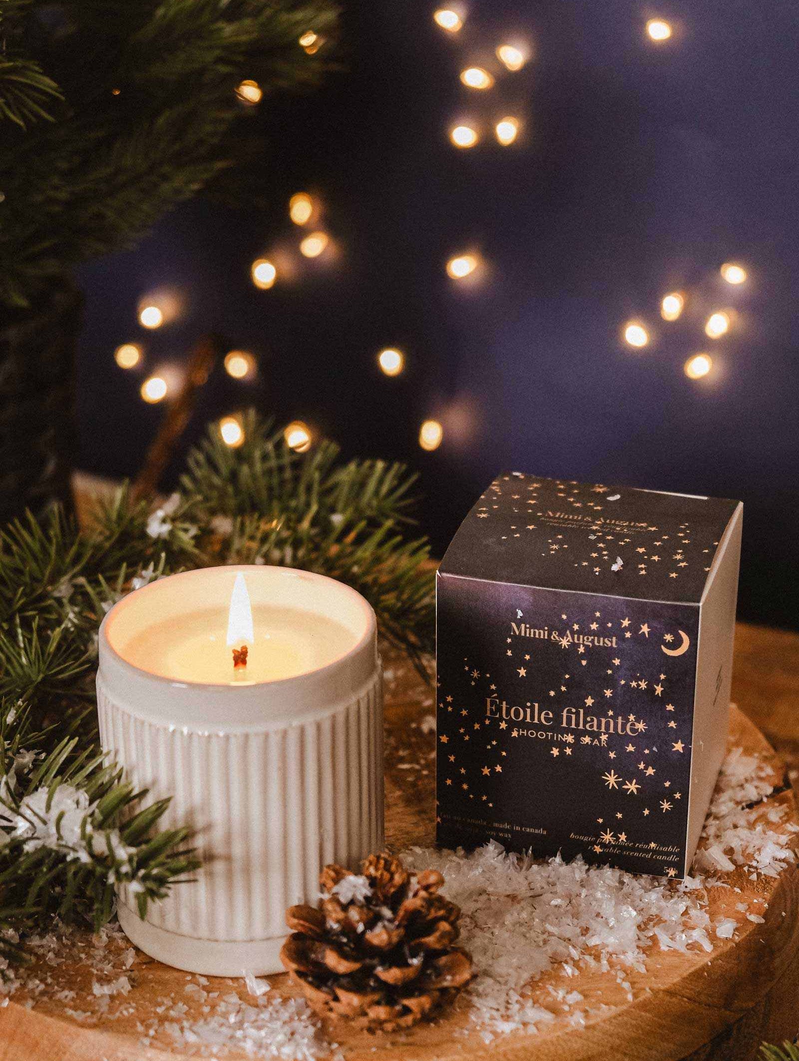 The Shooting Star - Christmas Candle by Mimi & August, in a ribbed container, sits next to a decorated box amid pine branches, a pinecone, and faux snow on wood; blurred lights evoke the cozy spirit of winter nights.