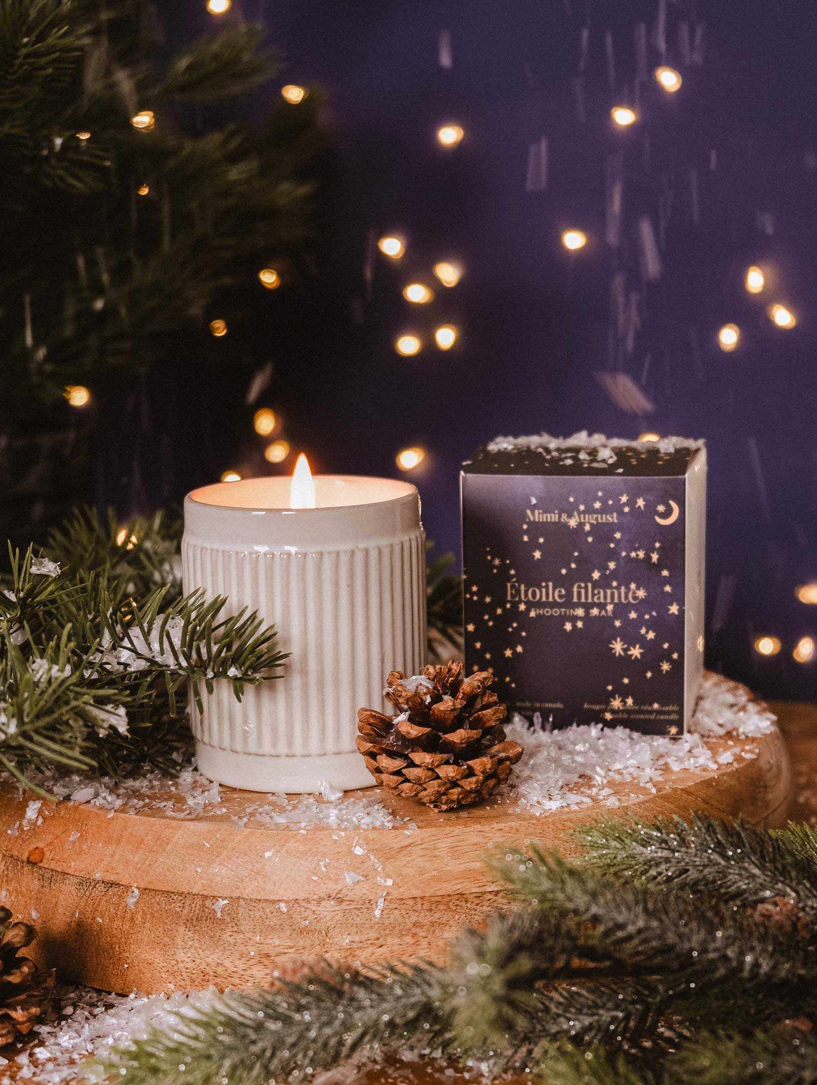 A Shooting Star - Christmas Candle by Mimi & August, a pine cone, and a festive box rest on a wooden surface with faux snow and pine branches, creating cozy holiday vibes with softly glowing lights in the background.