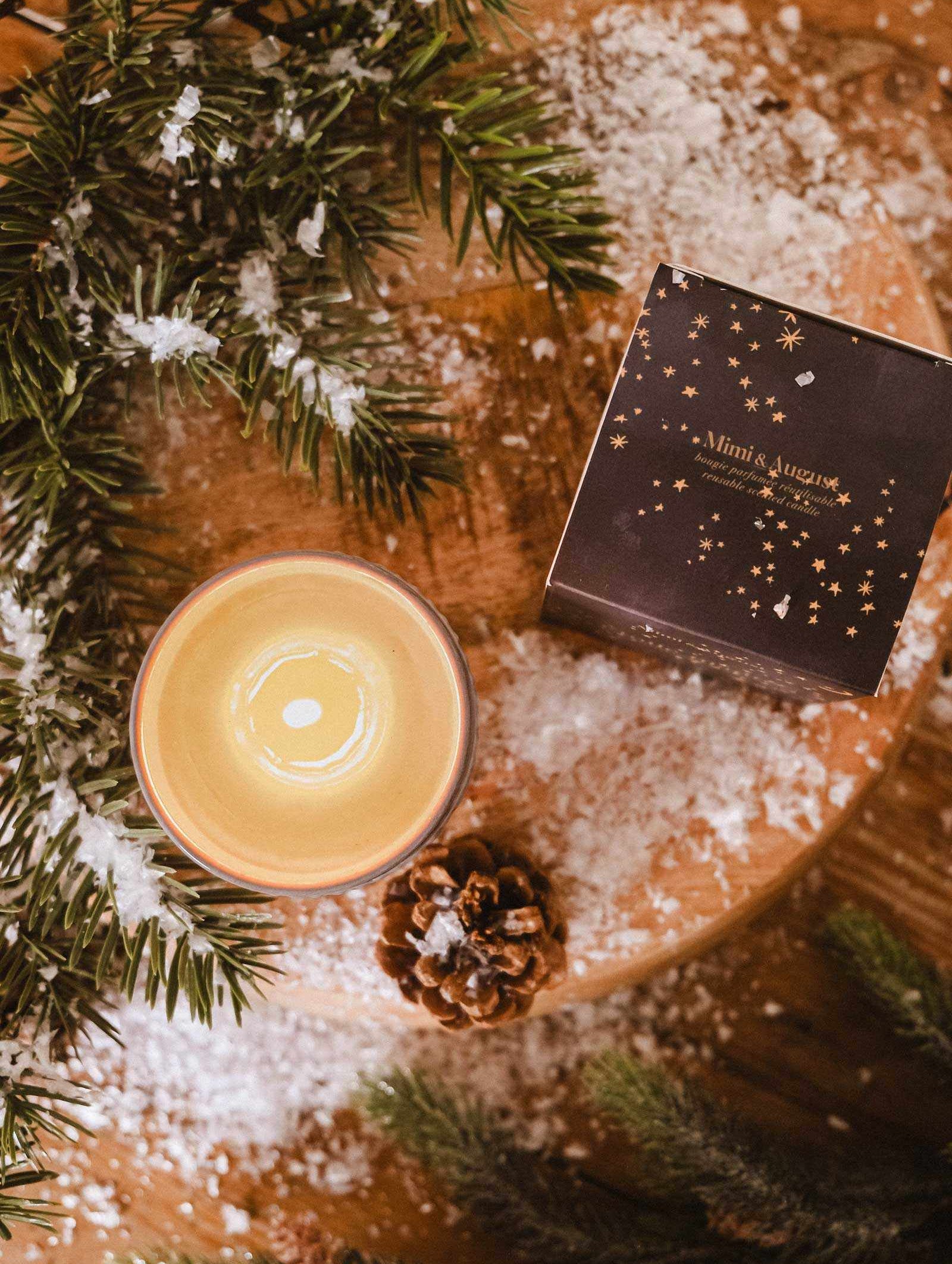 A Mimi & August Shooting Star Christmas Candle, pine cone, artificial snow, pine branches, and a dark starry gift box are arranged on a wooden surface.