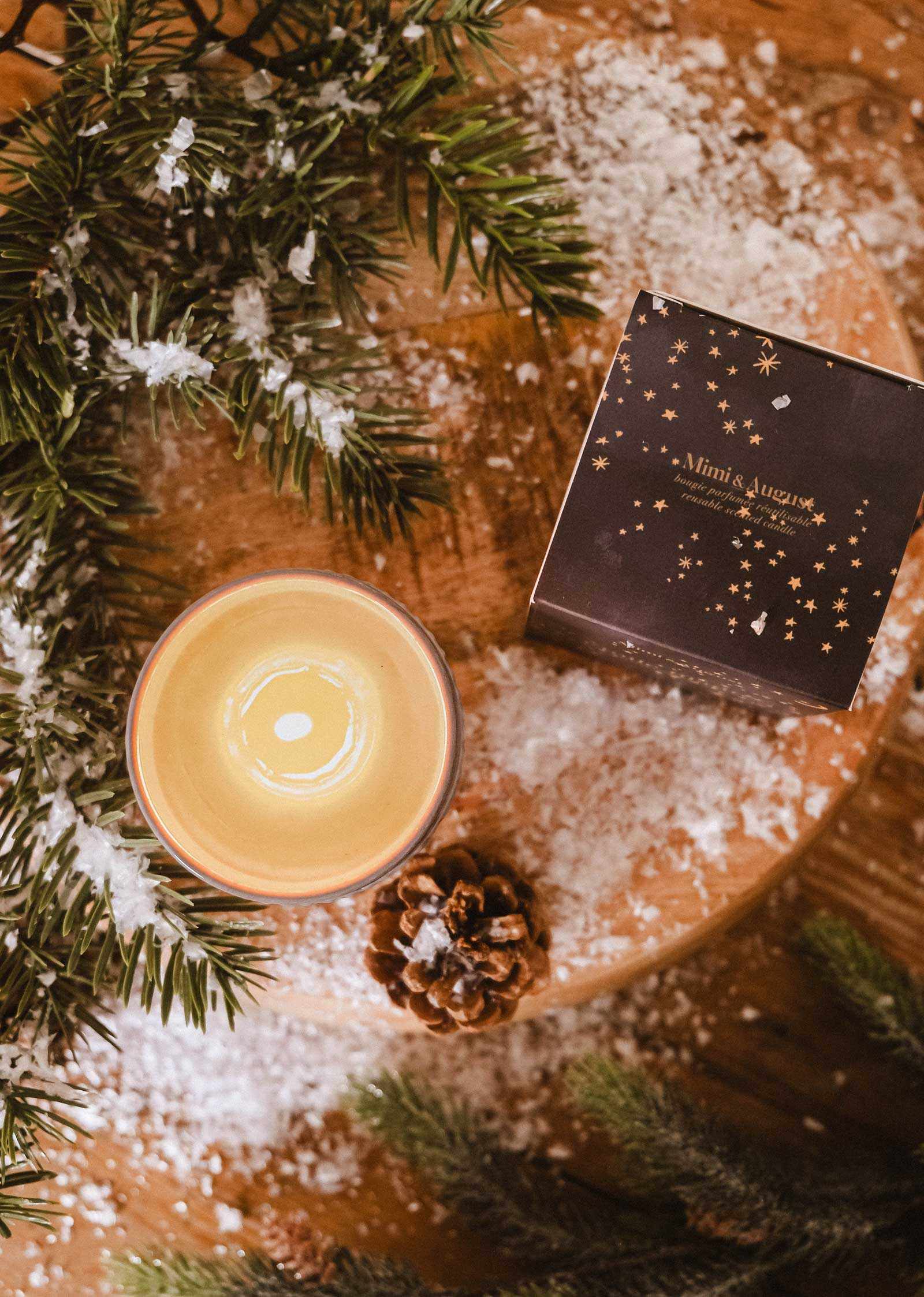 A Mimi & August Shooting Star Christmas Candle, pine cone, artificial snow, pine branches, and a dark starry gift box are arranged on a wooden surface.