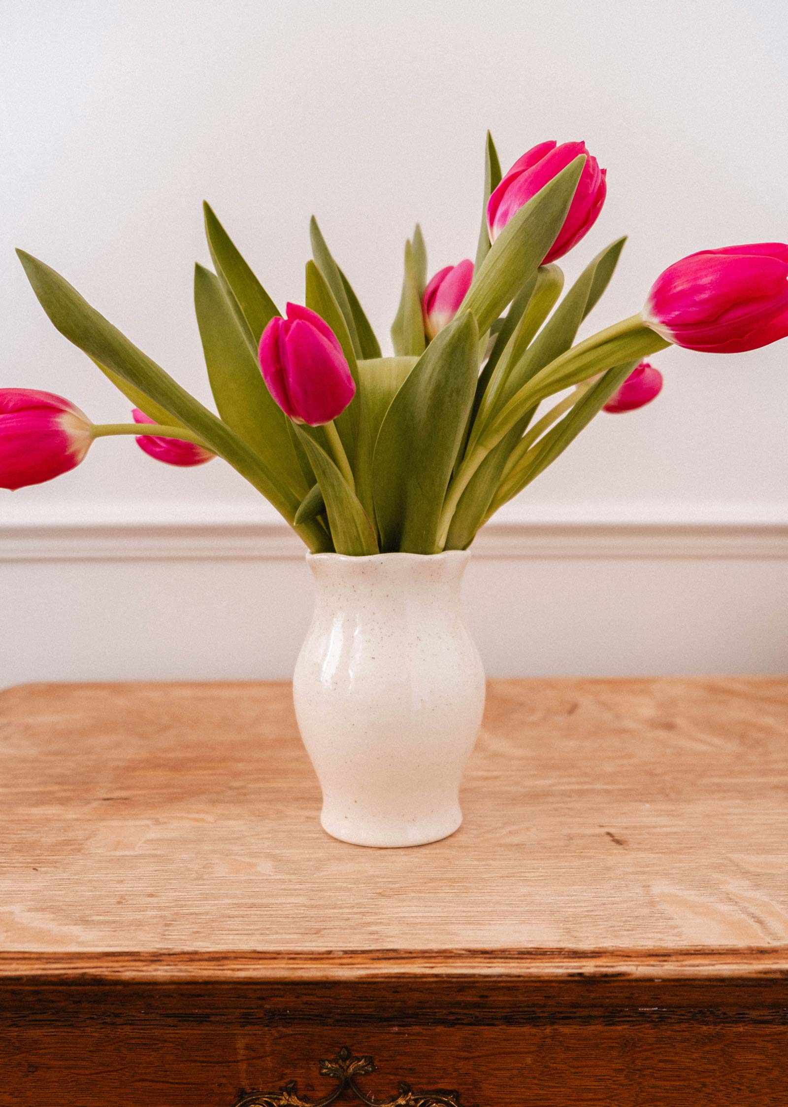 Le Vase en céramique Silhouette de Mimi & August, garni de tulipes roses, apporte du charme à votre décor sur une table en bois contre un mur blanc.