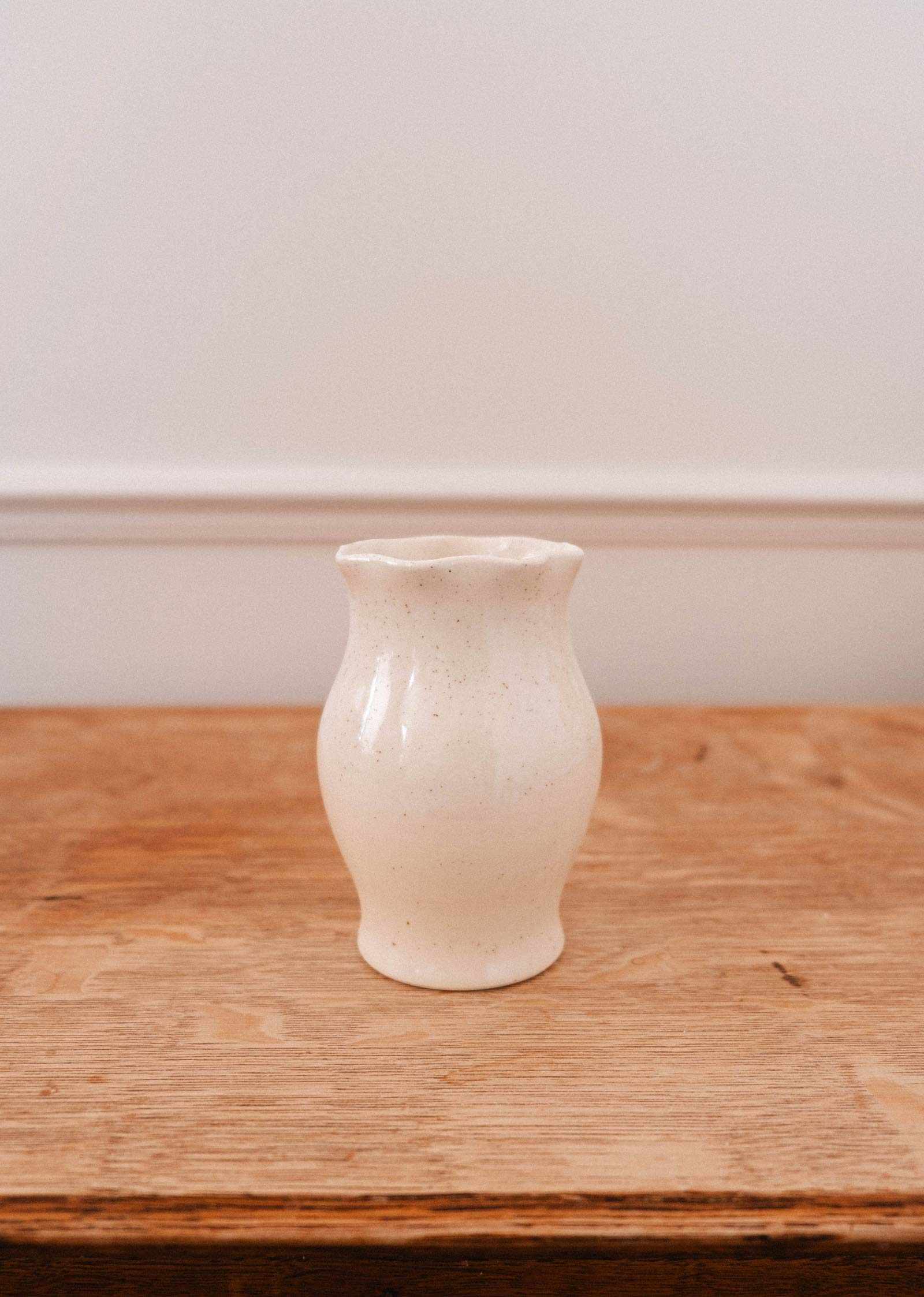 Le Vase en céramique Silhouette de Mimi & August, artisanal à la finition crème, repose sur une surface en bois contre un mur blanc — idéal pour ajouter une touche élégante à votre décor.