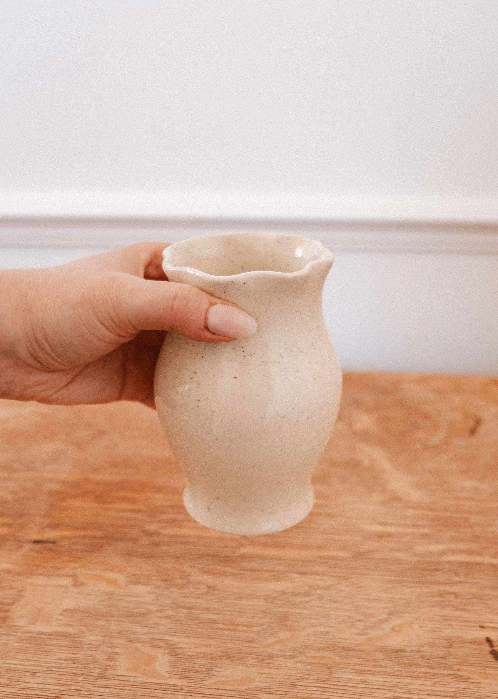 Une main tient le Vase en céramique Silhouette de Mimi & August, au rebord ondulé, au-dessus d'une surface en bois — un accent élégant pour tout décor.