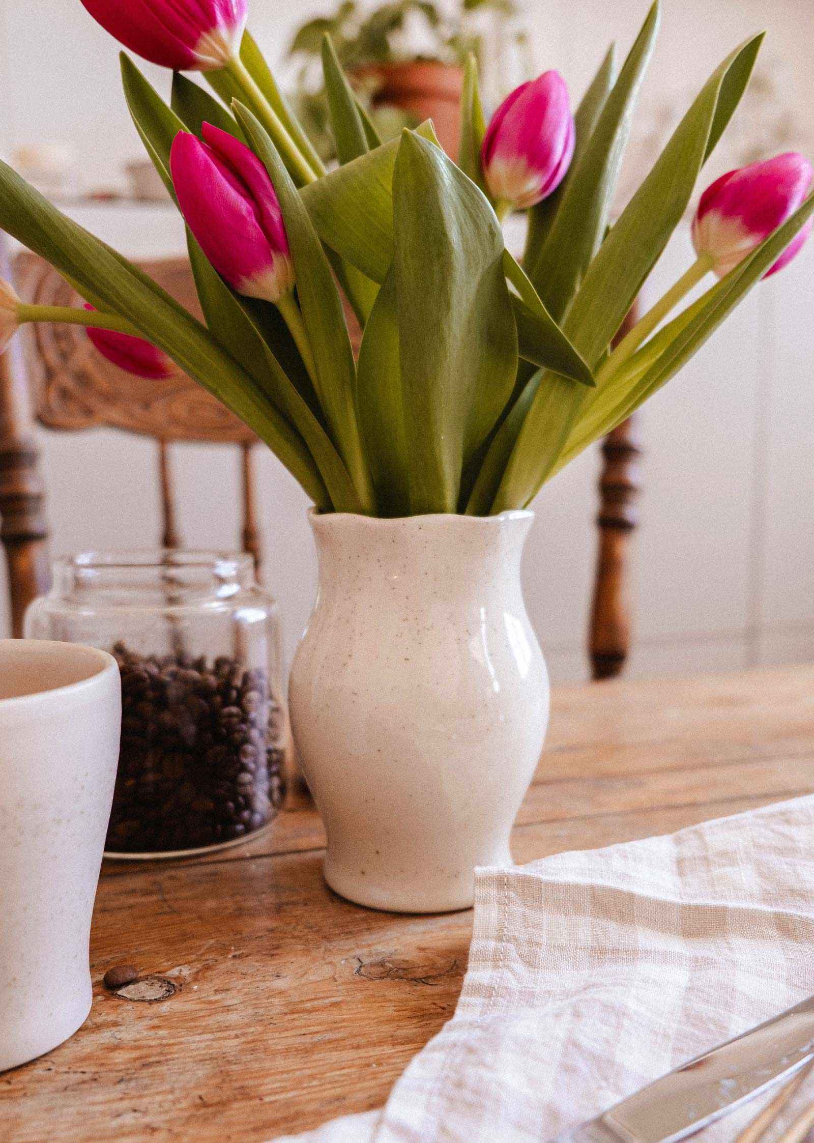 Le Vase en céramique Silhouette de Mimi & August, garni de tulipes roses, est présenté aux côtés d'une tasse, d'un pot de grains de café et d'une serviette sur une table en bois — ajoutant du charme à tout décor.