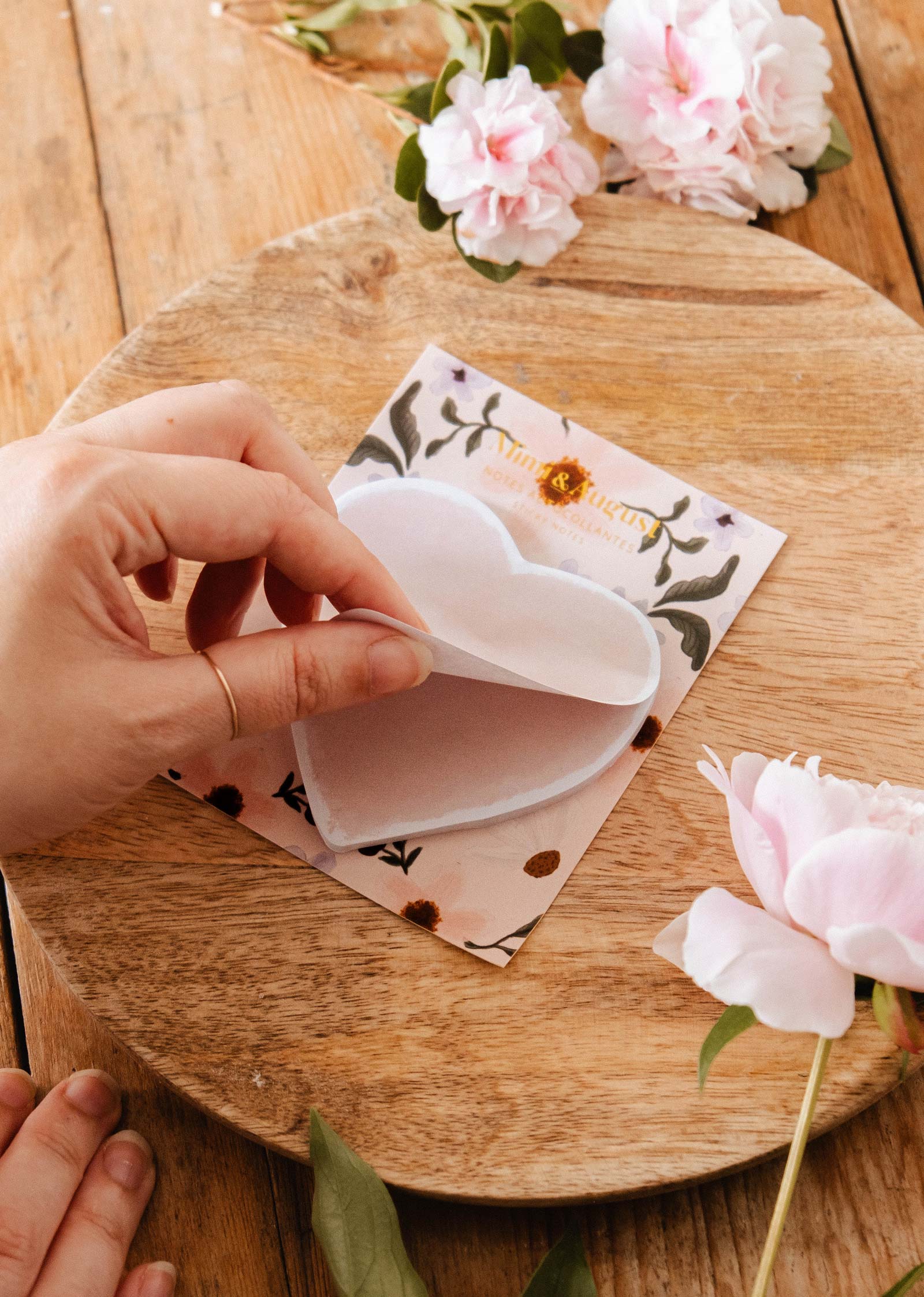 A hand peels a Mimi & August Soft Heart - Sticky Note, shaped like a heart, from a floral notecard on a wooden surface, with pink flowers nearby.